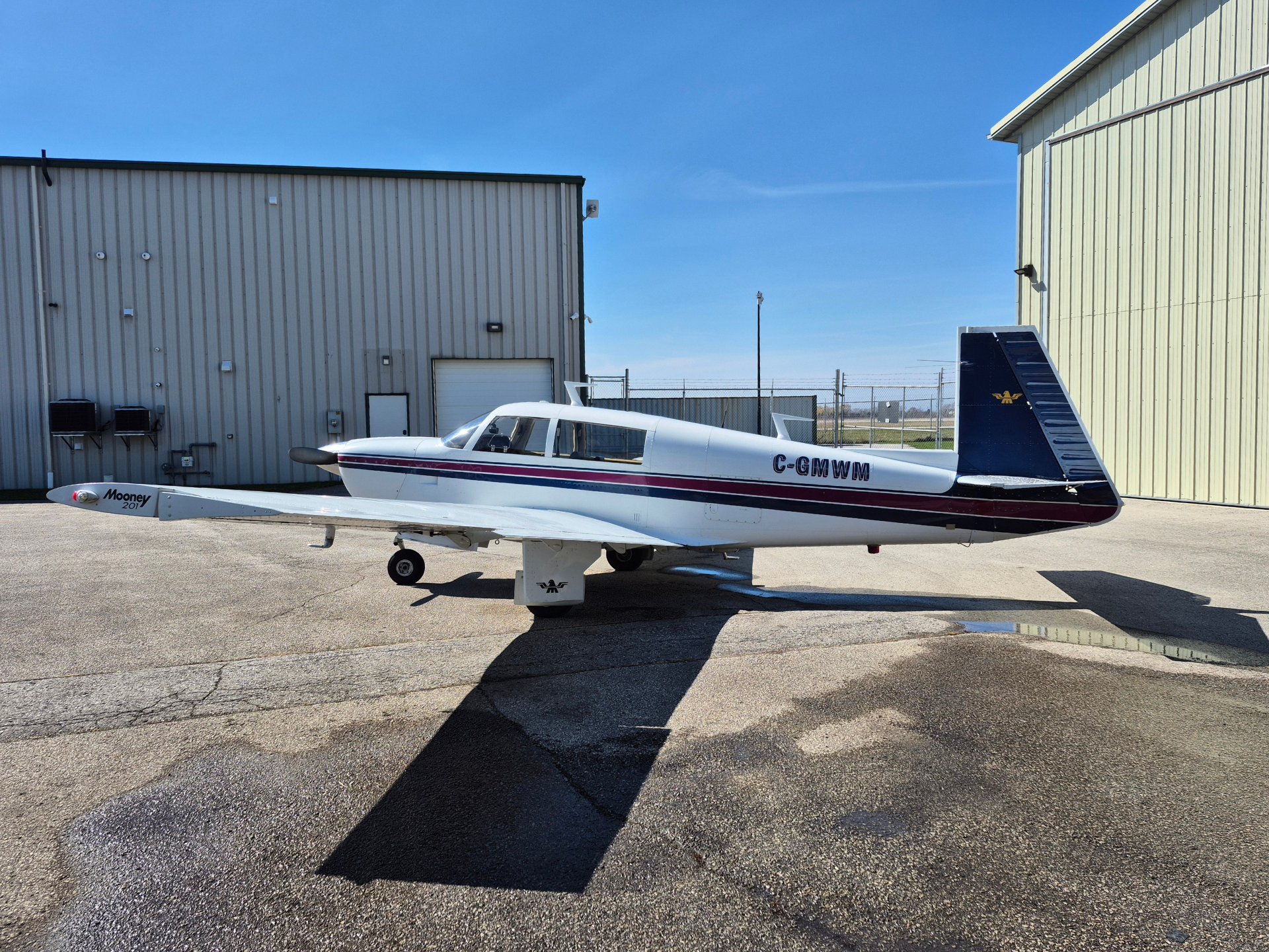White single-engine airplane on a cracked tarmac in front of beige hangars, clear blue sky above. Registration C-GMWM on fuselage, blue tail with logo.