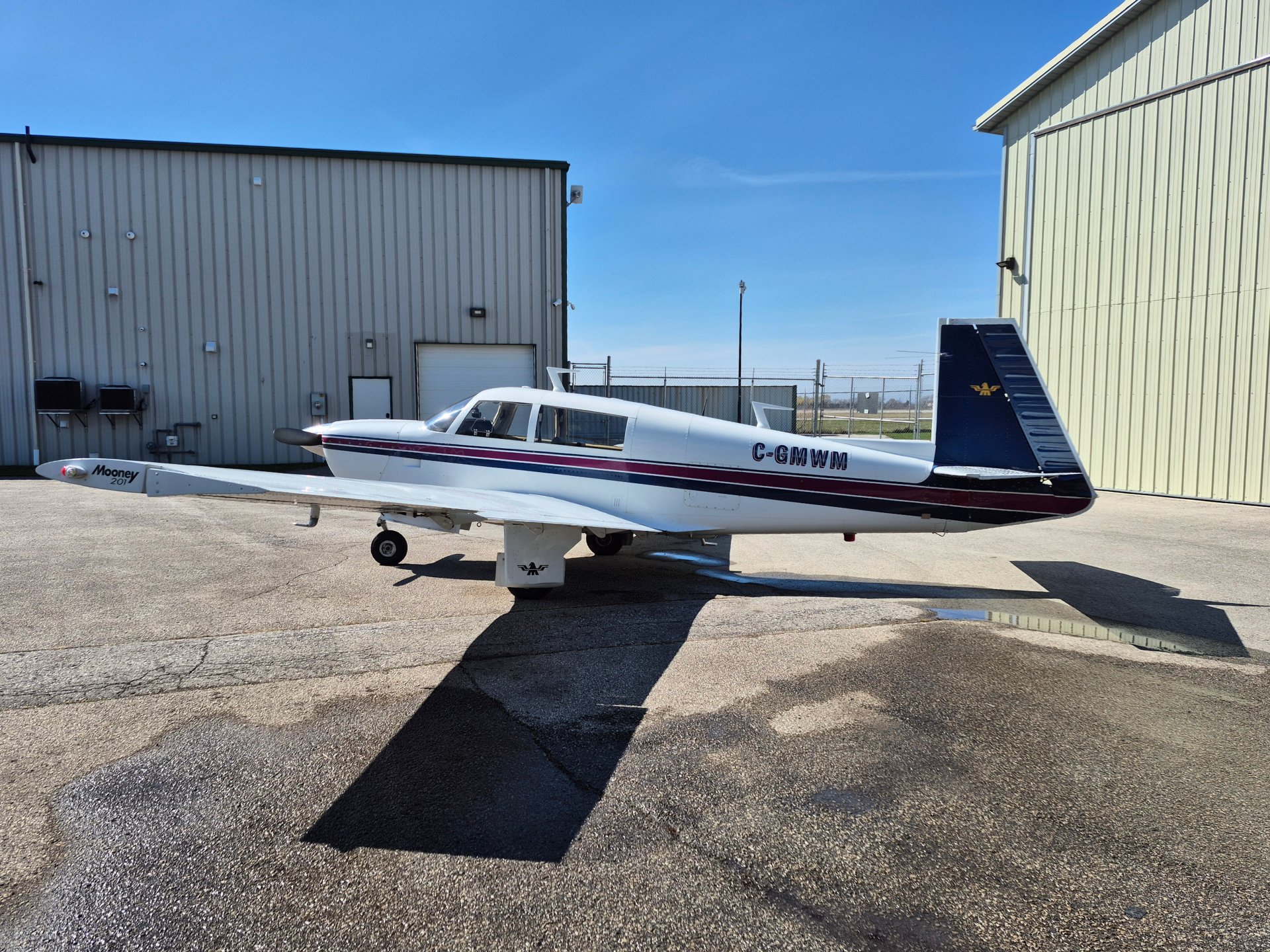 White single-engine airplane with red/blue stripe parked on tarmac beside hangars under a clear blue sky; registration C-GWMM visible on fuselage.