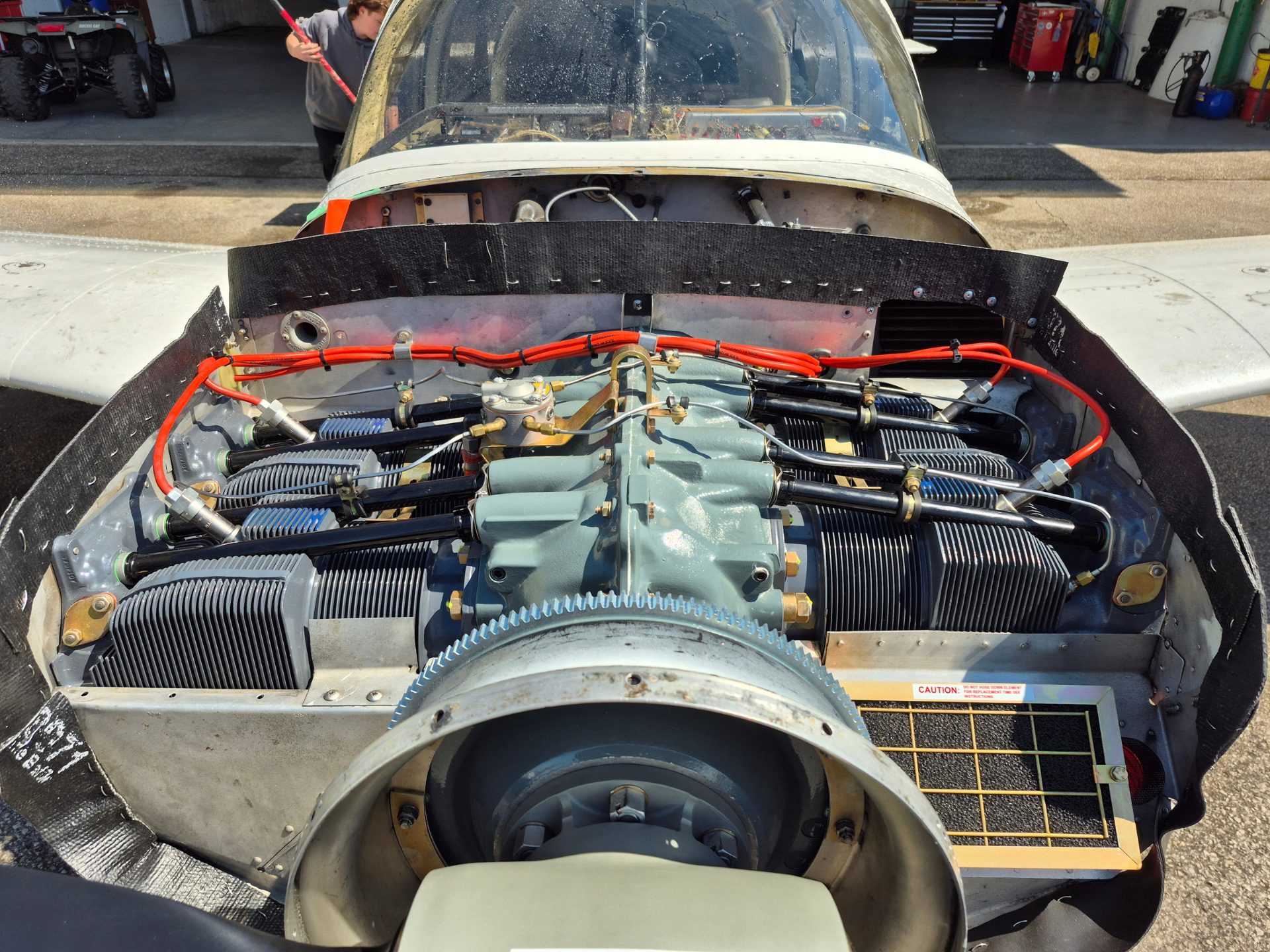 Front view of an aircraft engine with exposed cylinders and bright red wiring, inside an open cowling in a hangar workshop