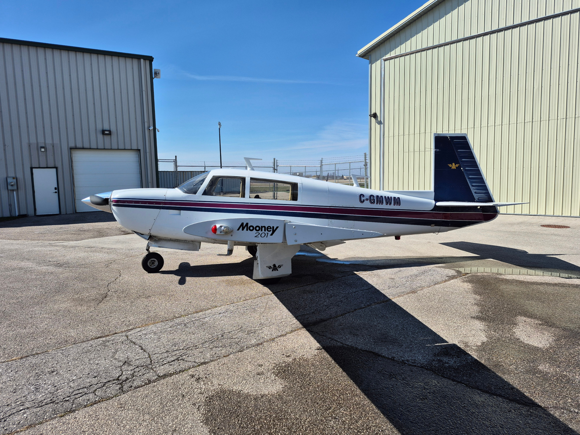 White Mooney 201 airplane with red/blue stripe parked on a sunlit tarmac beside metal hangars; registration C-GMWM visible on fuselage