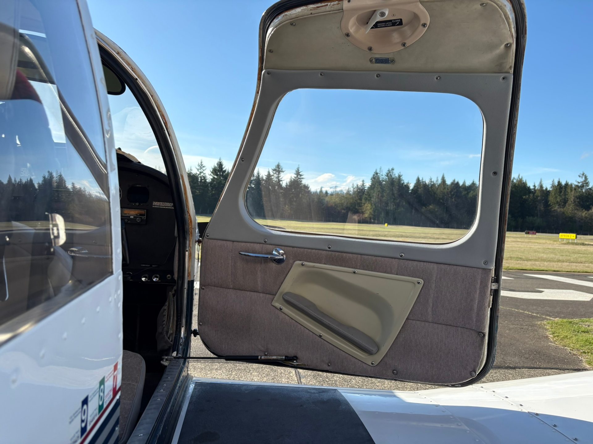 Open airplane door showing the interior door panel with fabric, window cutout, and a clear blue sky with runway and trees outside.
