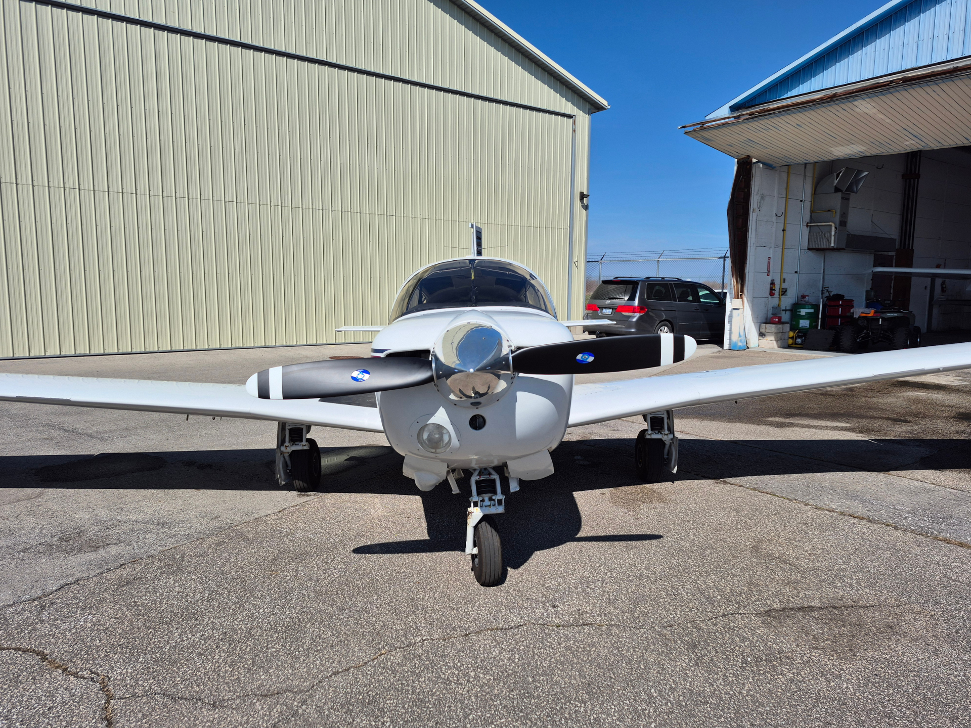 Front view of a small white propeller airplane parked on a tarmac in front of a pale-green hangar under a blue sky.