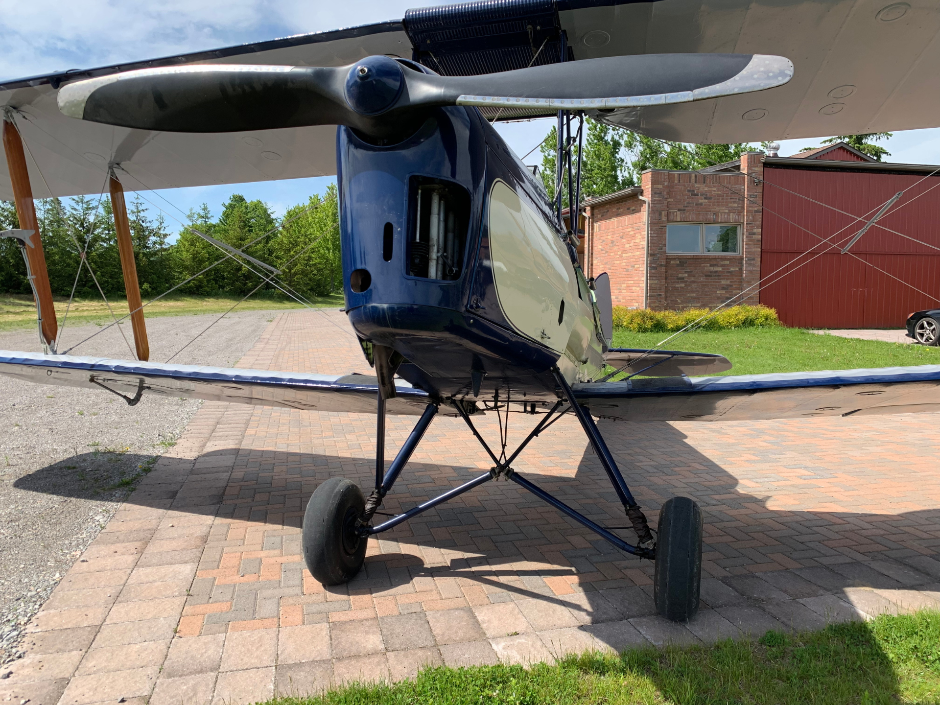 Blue vintage biplane with propeller-facing nose, parked on a brick surface with a red brick hangar in the background and grassy area nearby.