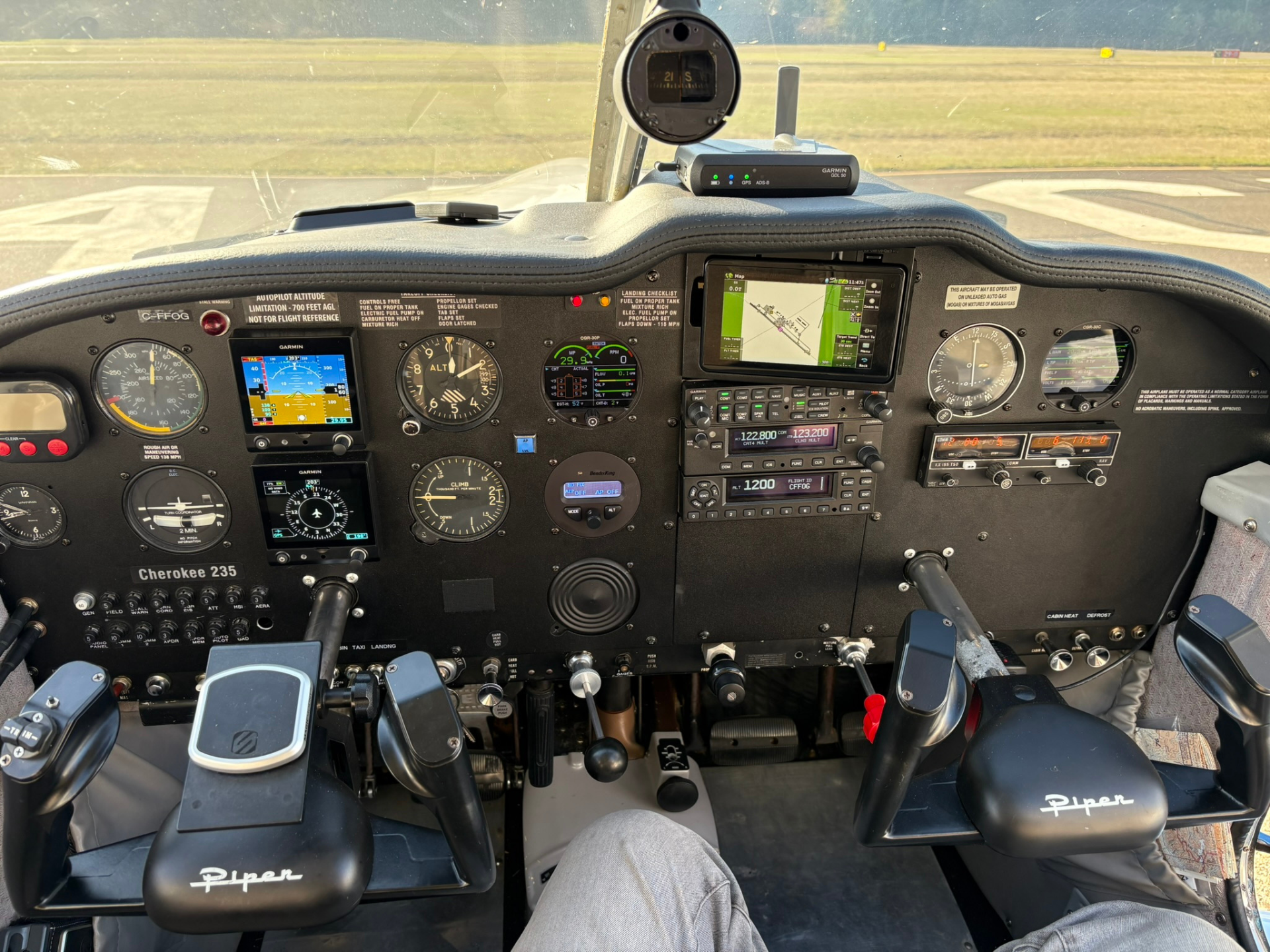 Cockpit of a small Piper Cherokee 235 with avionics, gauges, and yokes visible, runway outside the windshield, pilot's knees in view.