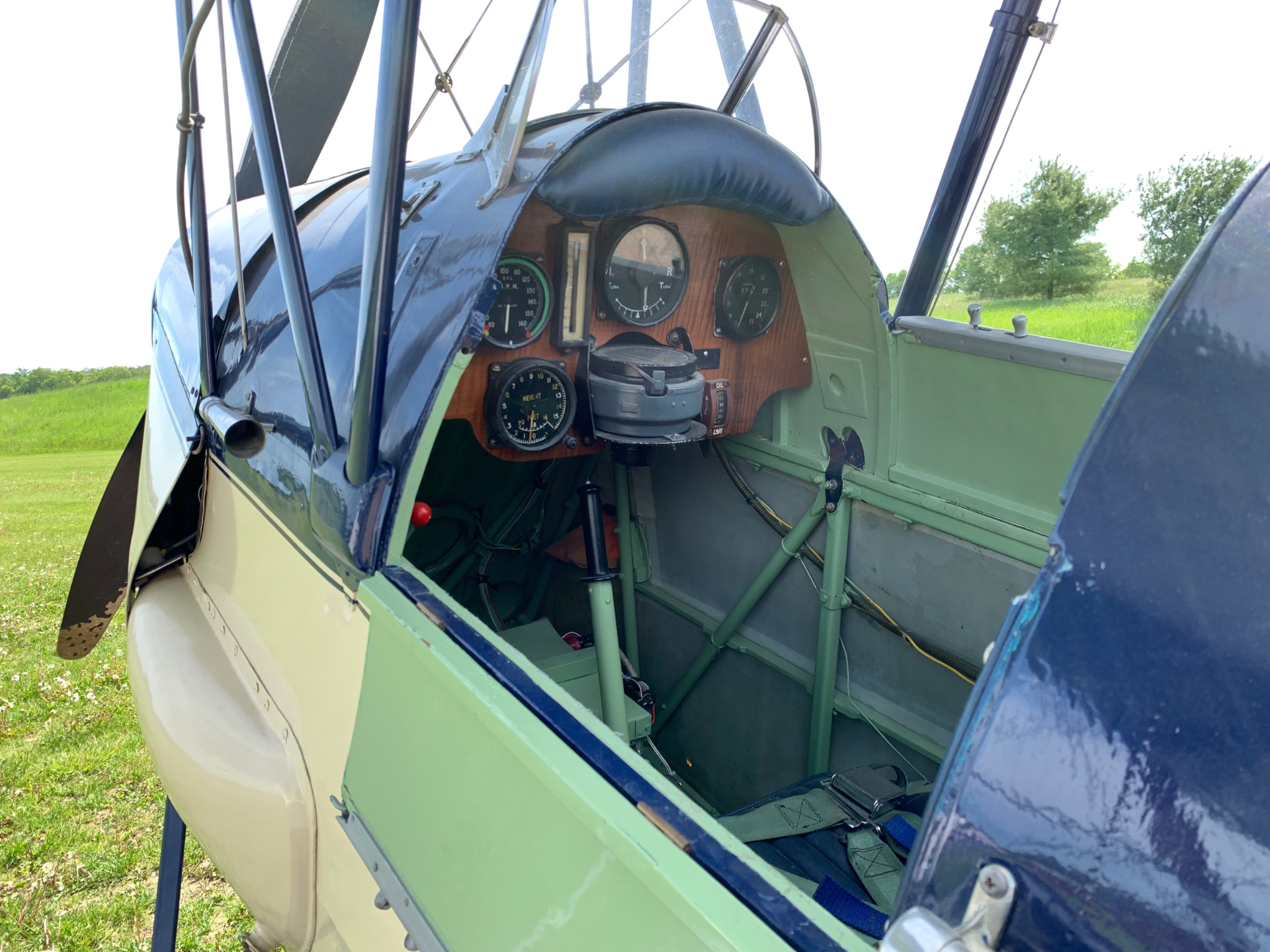 Open cockpit of a vintage biplane showing a wooden instrument panel with round gauges and a central compass, against a green field outside.