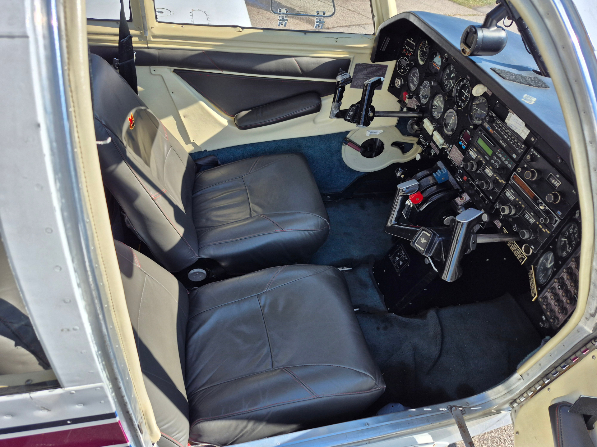 Interior of a small airplane cockpit with two gray leather seats and a complex instrument panel on the right, including yokes, dials, and switches.