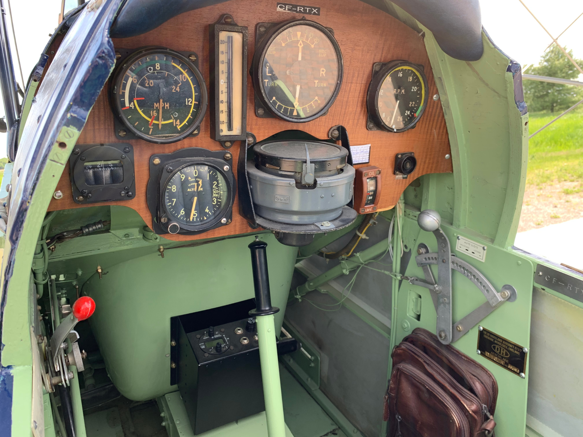 Vintage aircraft cockpit with a wooden instrument panel featuring multiple round gauges, including speed/altitude indicators, a central large dial, and a leather flight bag nearby.