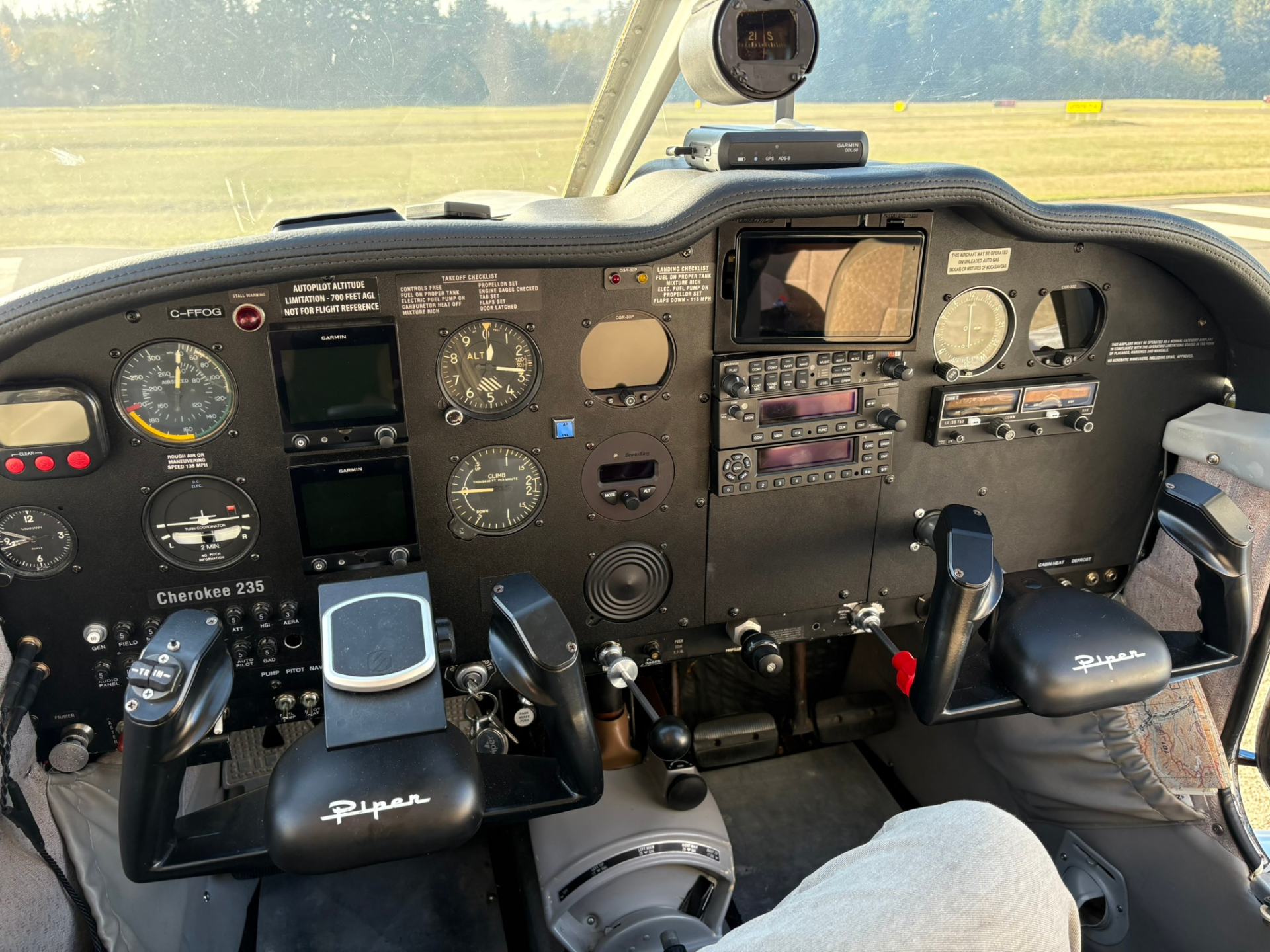Cockpit of a Piper Cherokee 235 with dual yokes, gauges, and avionics panel visible. Outside field seen through windshield.