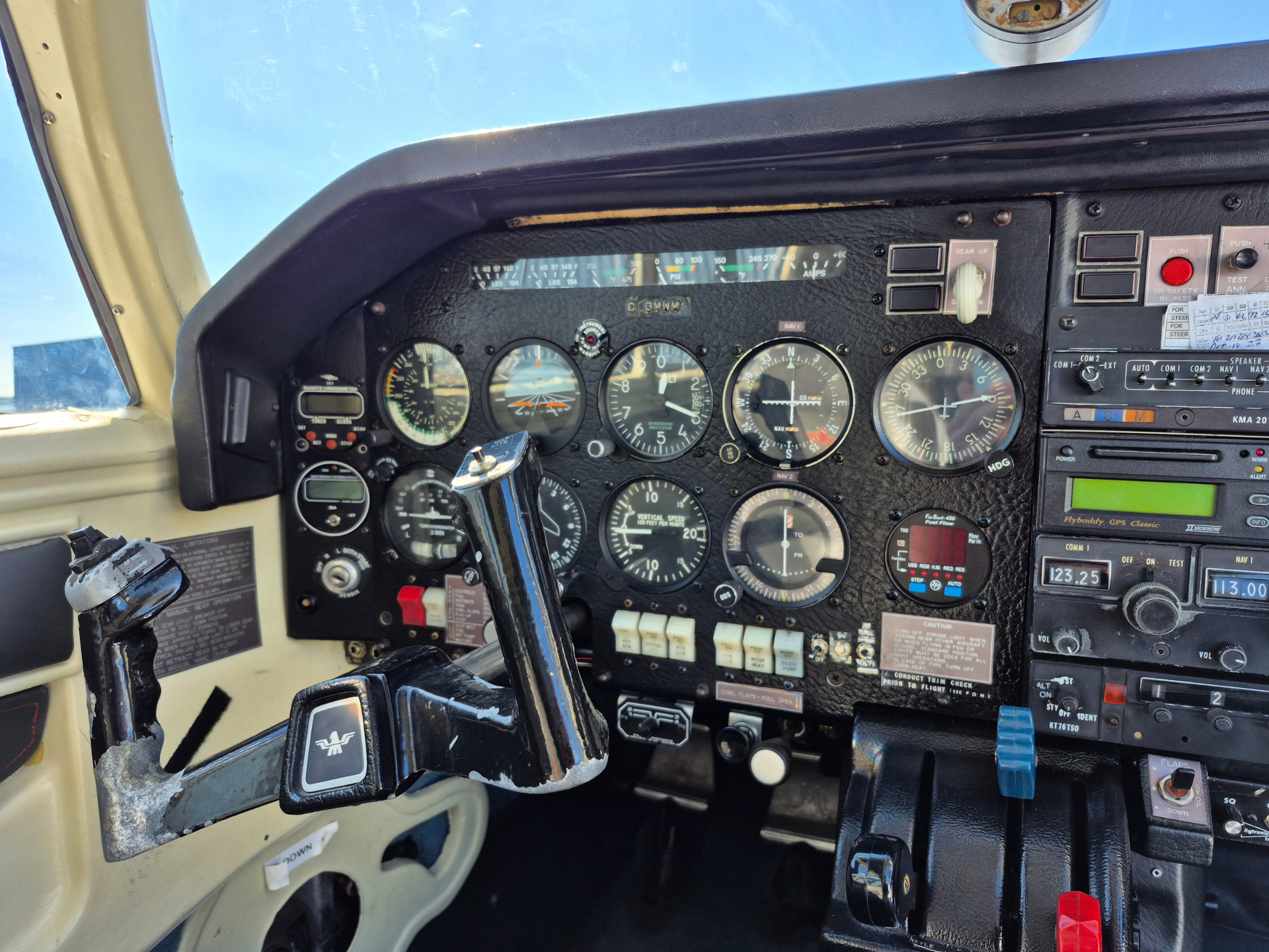 Close-up of a small aircraft cockpit with a dense array of analog gauges, flight yoke, and avionics panels showing blue sky outside the windshield