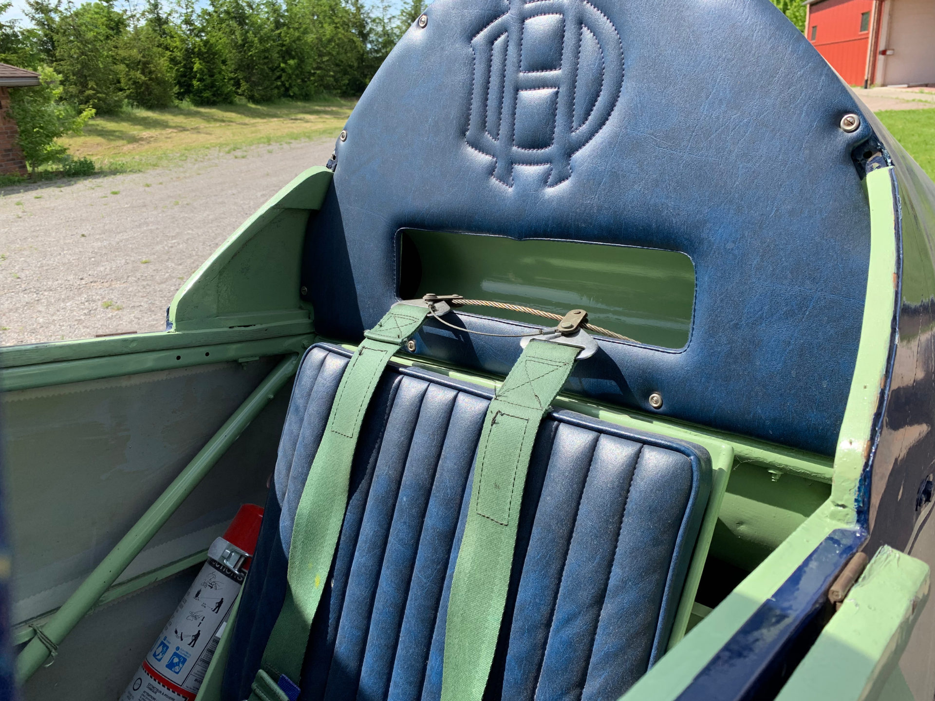 Close-up of a vintage airplane cockpit seat with blue padding and green straps, exterior green fuselage visible outside the window area.