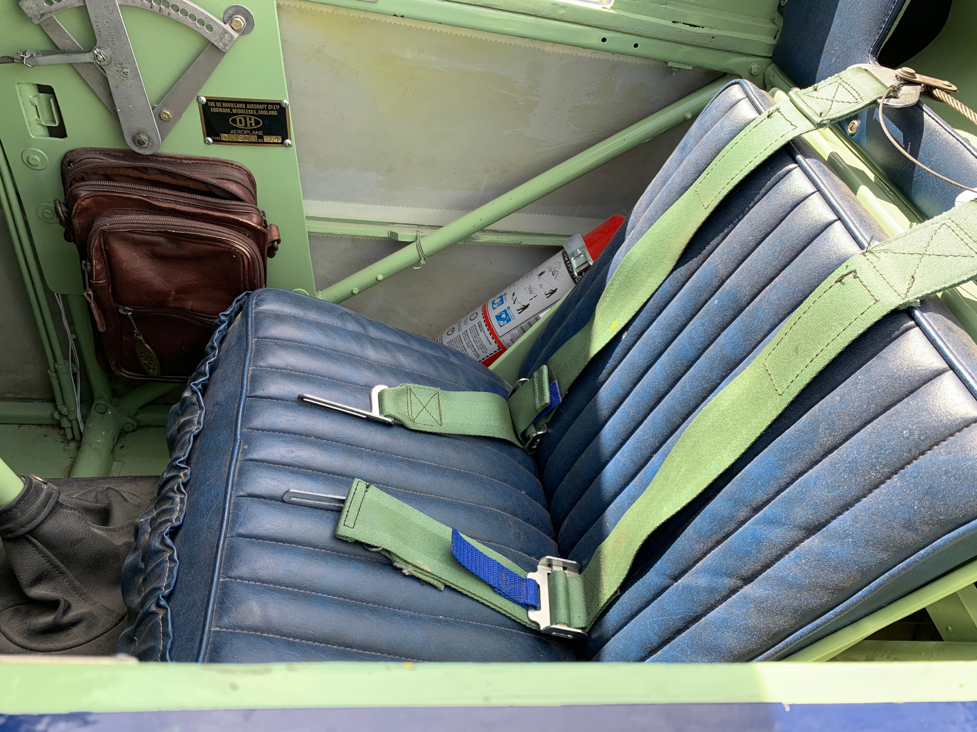 Inside a vintage light-green aircraft cabin with blue cushioned seats and green seat belts, brown leather bag tucked nearby.
