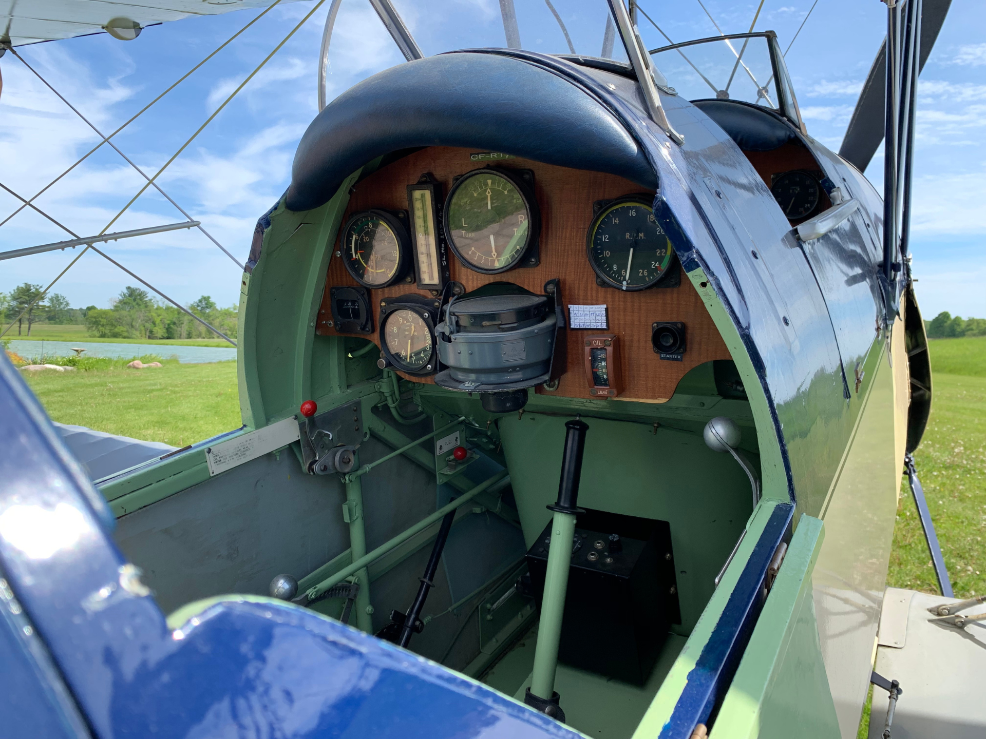 Cockpit of a vintage biplane with wooden instrument panel, showing multiple analog gauges and a central compass, surrounded by green and blue fuselage under a clear sky.