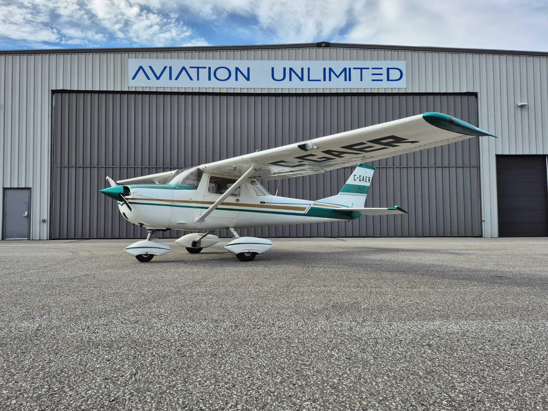 Small single-engine floatplane in front of a gray hangar with a large 'AVIATION UNLIMITED' sign above it, teal accents on nose and tail, landing gear visible.