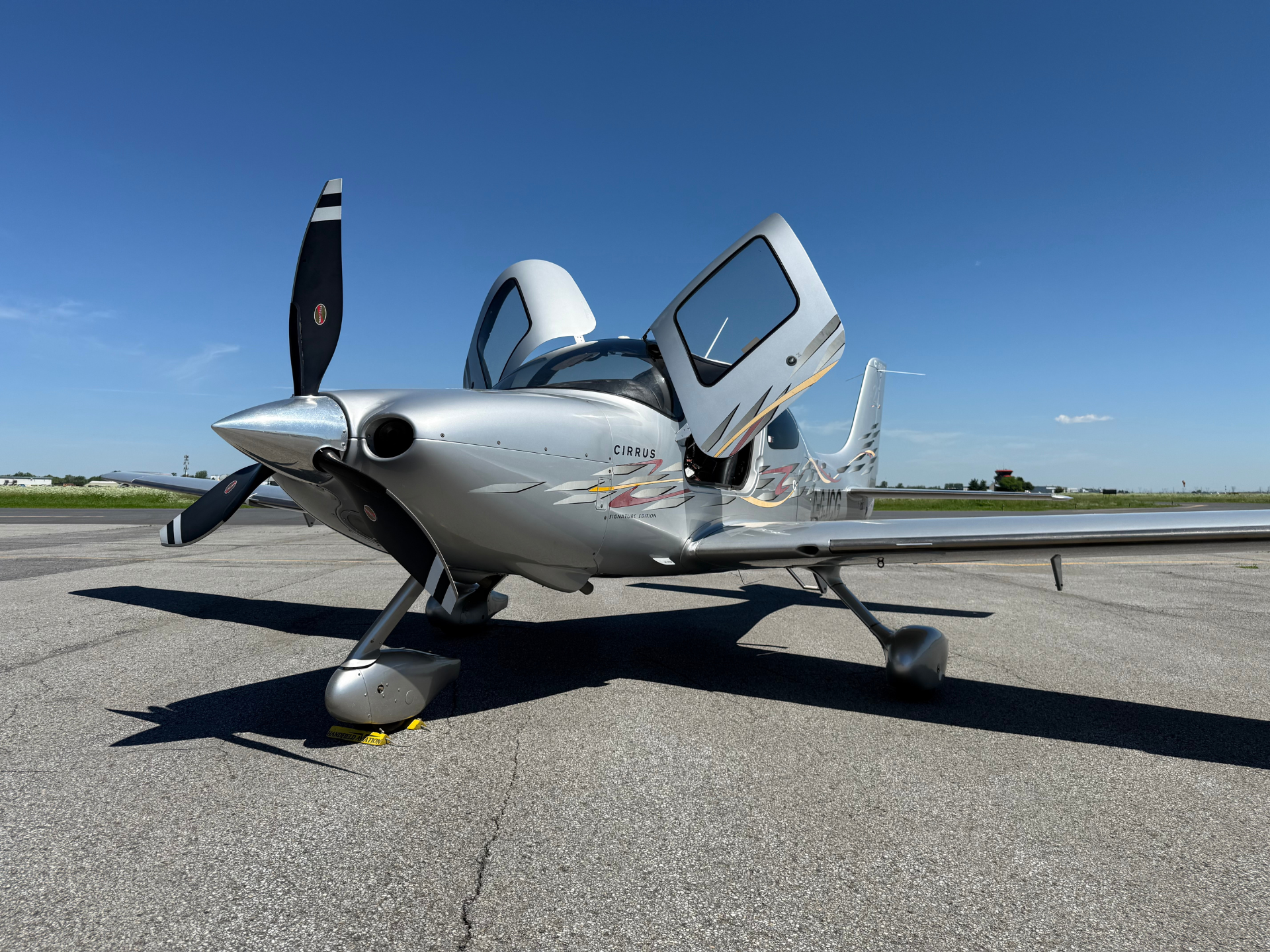 Silver Cirrus aircraft on a sunny tarmac with doors open and propeller visible, ready for boarding at a small airfield
