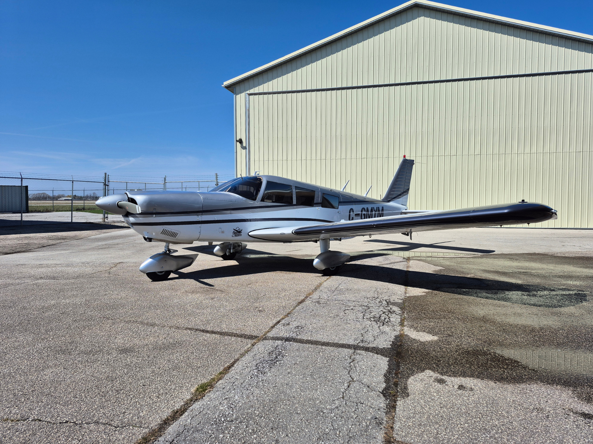 Small silver single-engine airplane parked on a cracked tarmac in front of a pale hangar under a clear blue sky
