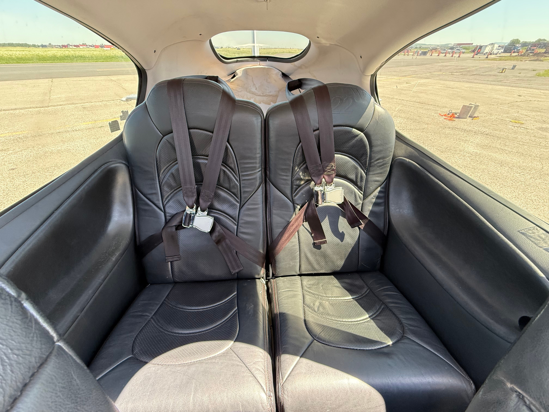 Two black leather aircraft seats with brown harnesses in a small airplane, view of runway outside through the side windows.