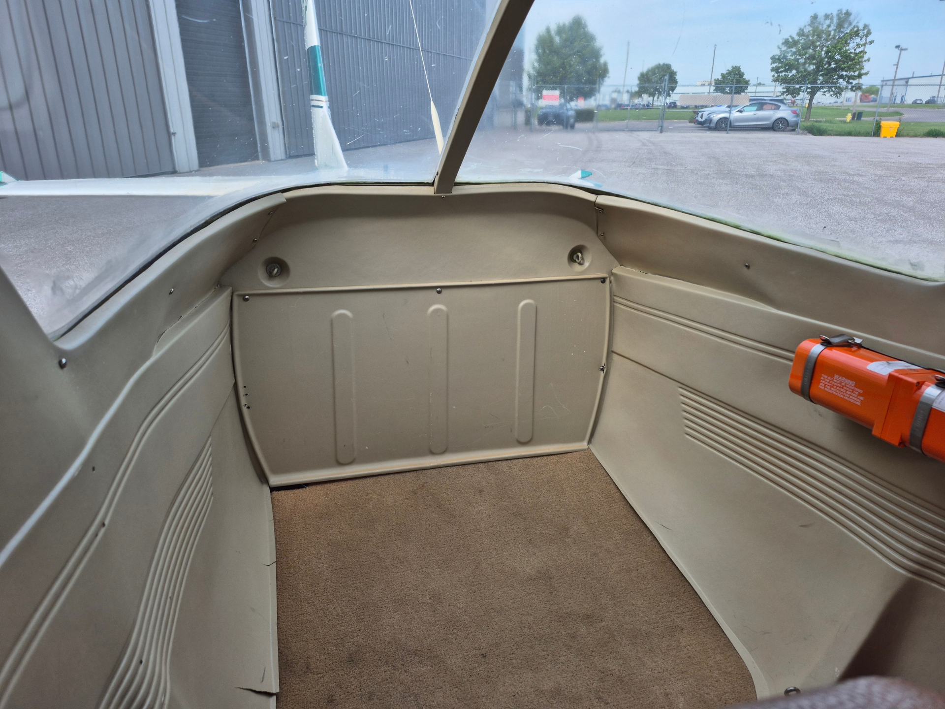 Interior of a small airplane cockpit with beige panels and brown carpet, looking out the windshield at a parking lot and hangar outside.