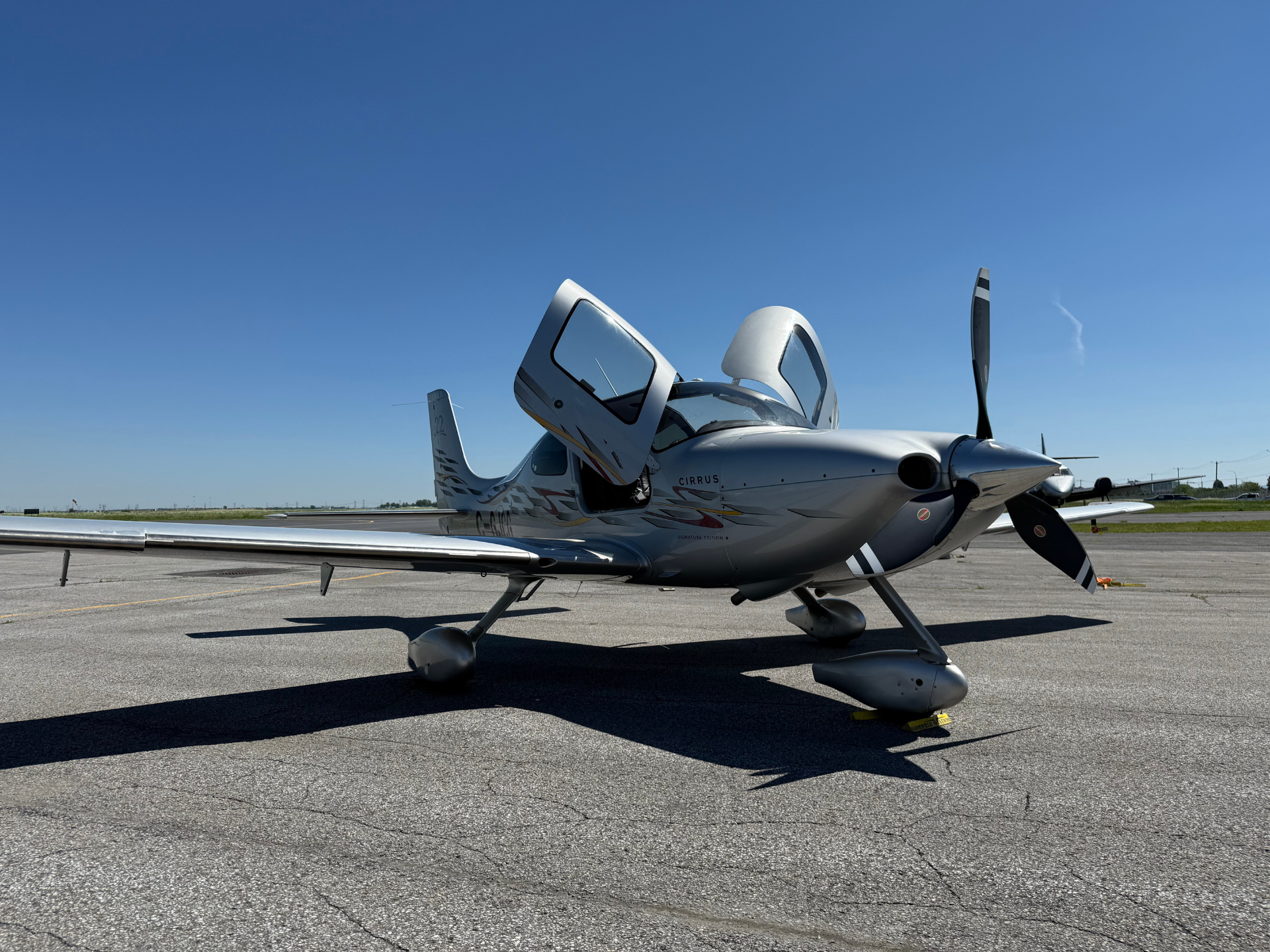 Silver Cirrus twin-engine aircraft with open passenger door on a sunny tarmac.