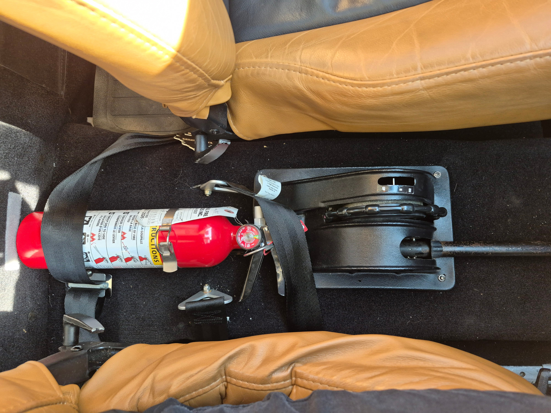 Fire extinguisher strapped to the car floor near the front seat mounting hardware inside a tan-leather interior.