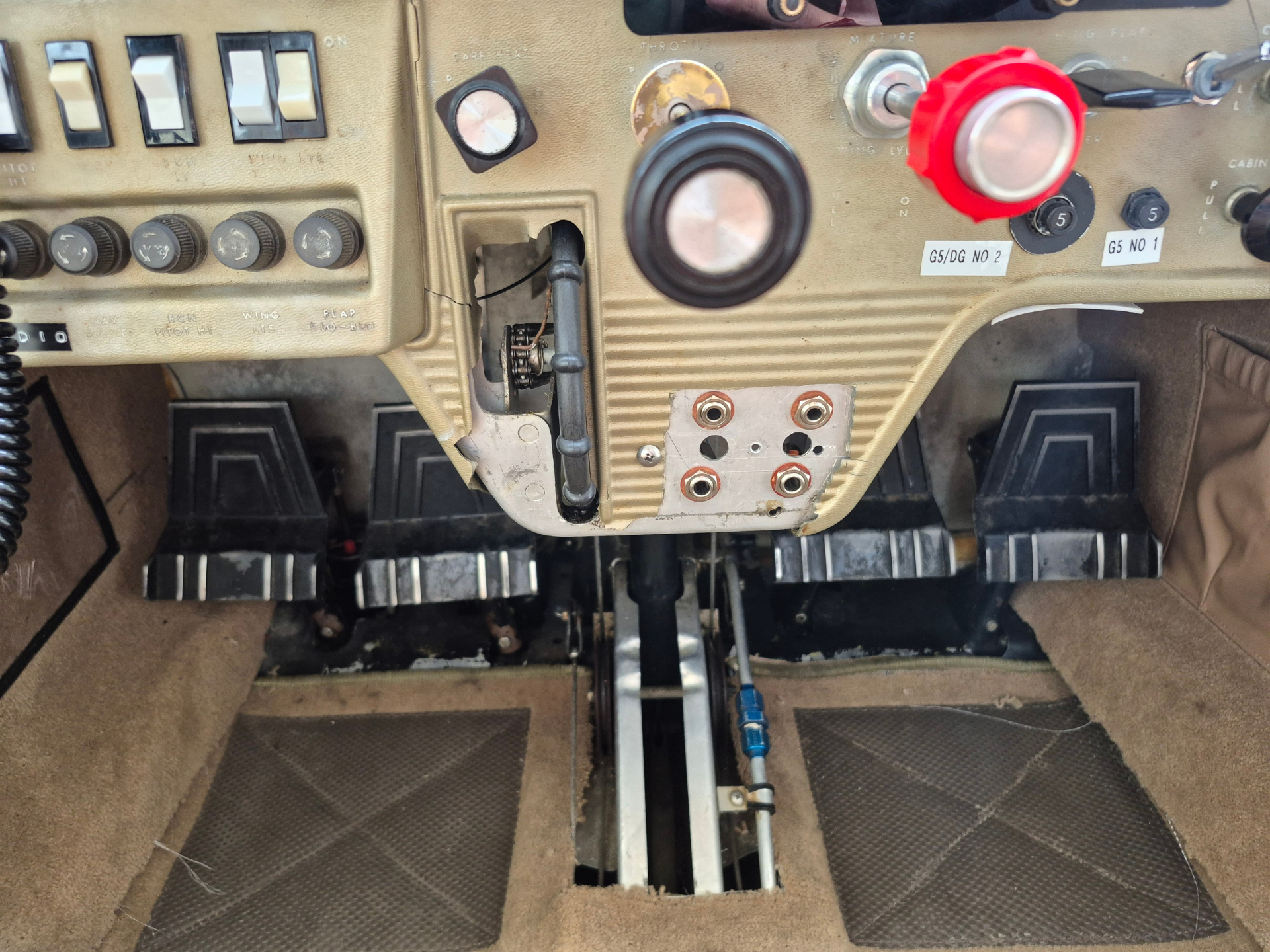 Cockpit center console with beige panels, multiple switches and knobs, and an open access panel showing internal mechanism.