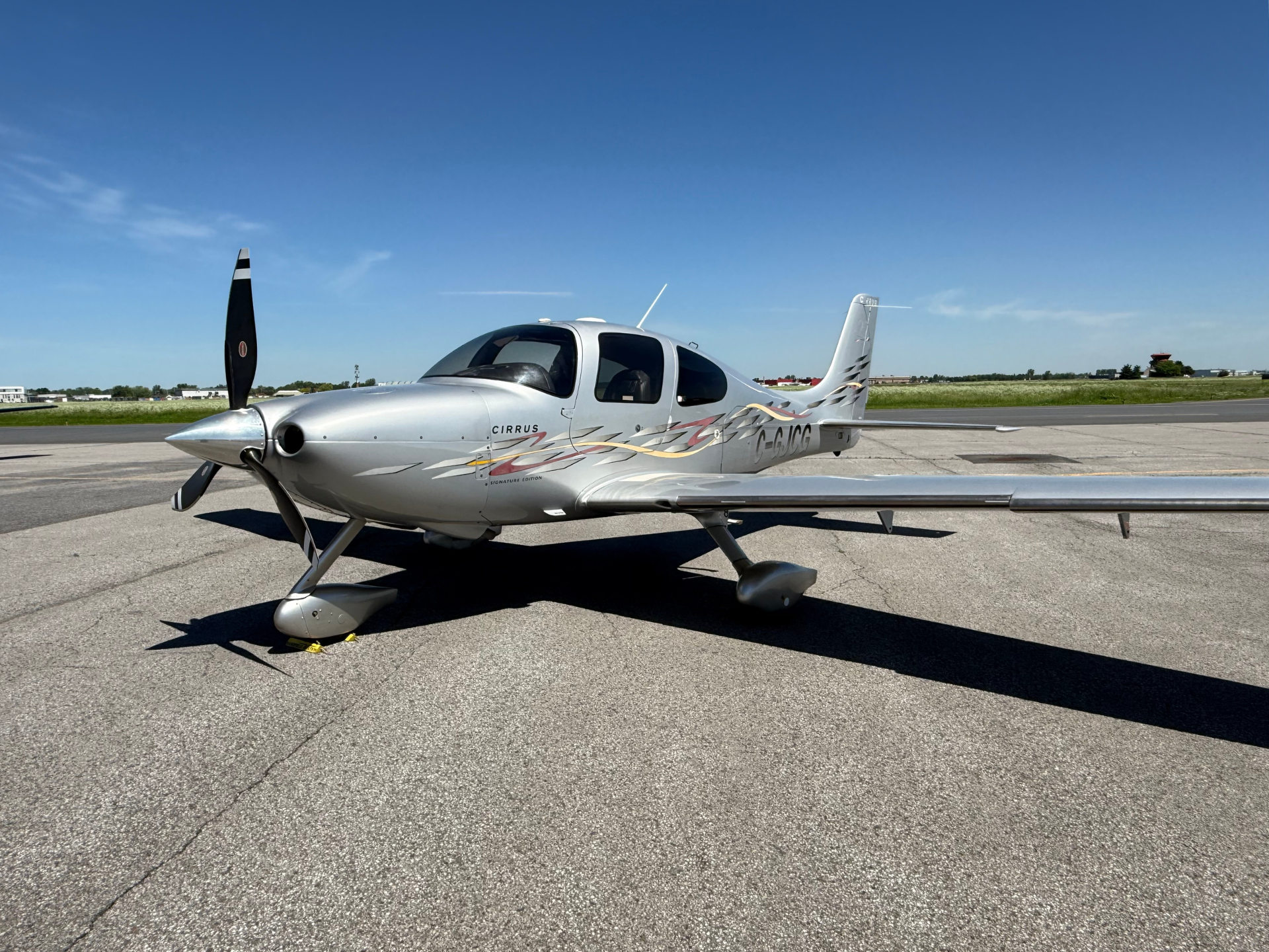 Silver Cirrus single-engine airplane on a tarmac, nose propeller visible, under a clear blue sky.