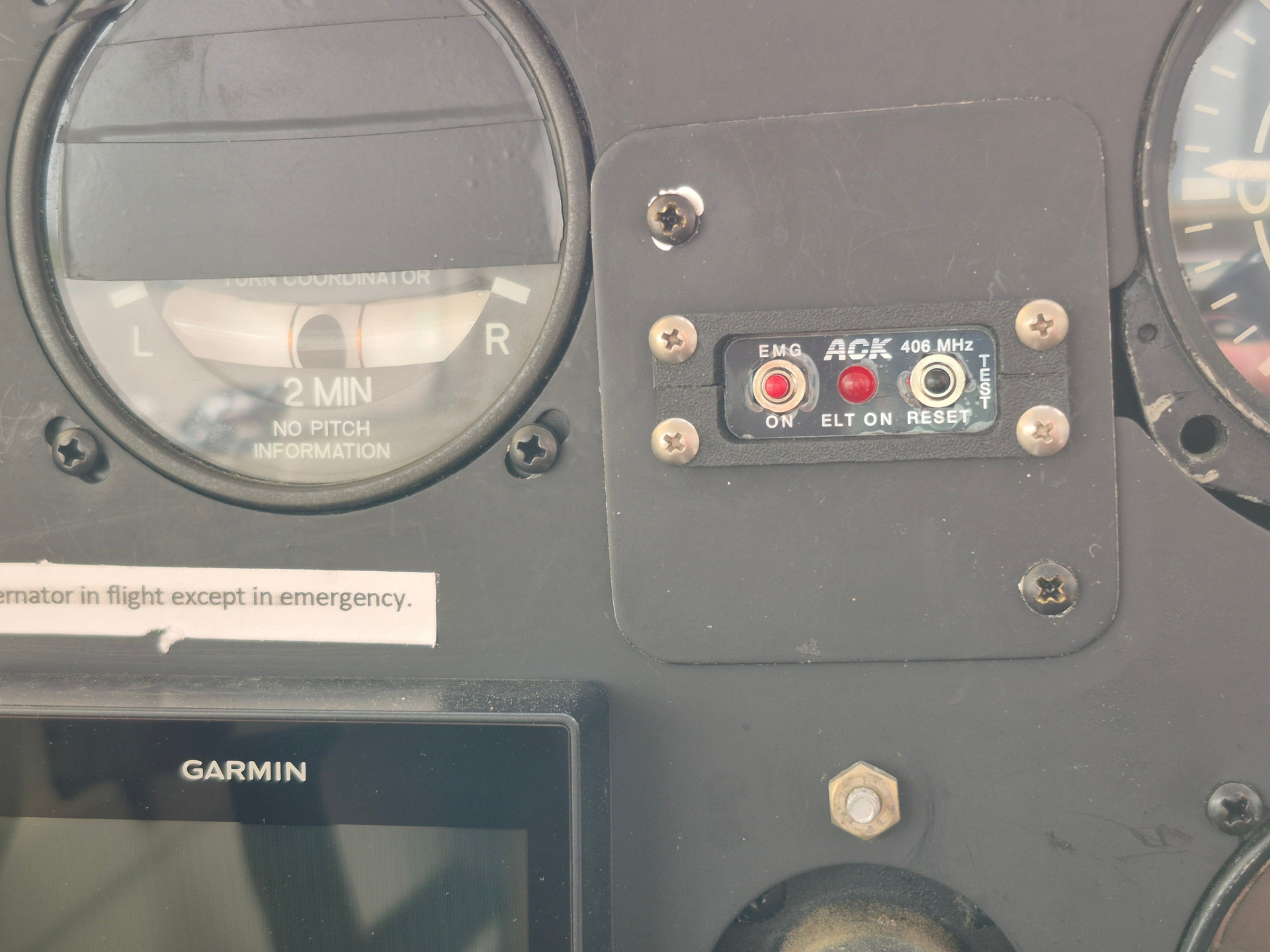 Close-up of an airplane cockpit panel: turn coordinator gauge on the left and a 406 MHz ELT control panel with EMG/ACK/TEST labels in the center, plus a Garmin screen below.