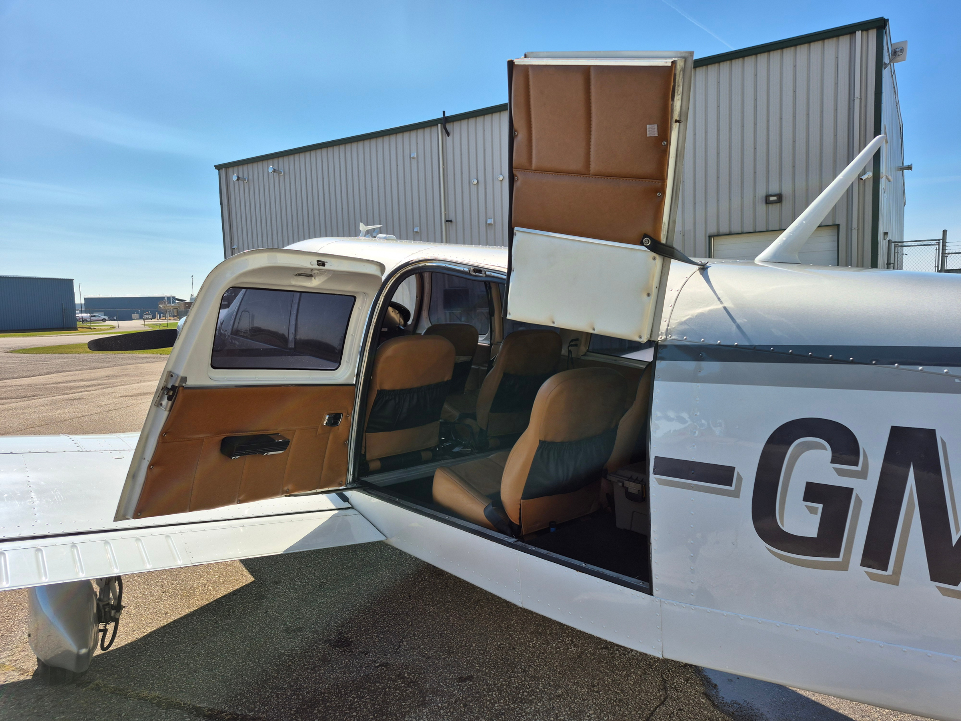 Small white airplane with open passenger door showing tan leather seats, parked on tarmac with a hangar in the background.