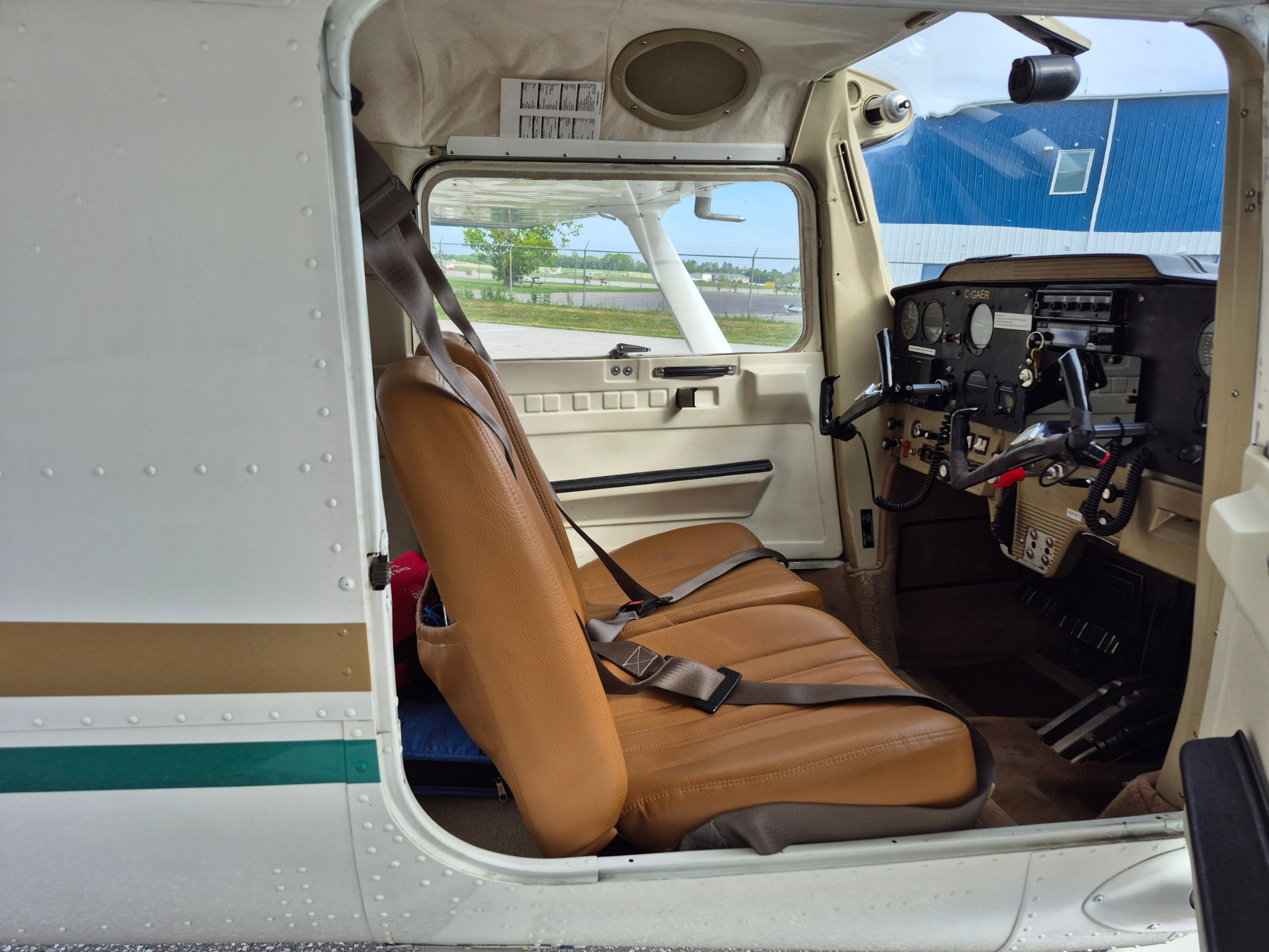 Interior of a small airplane cockpit with tan seats and instrument panel, open side door; runway and a blue hangar are visible through the window outside.