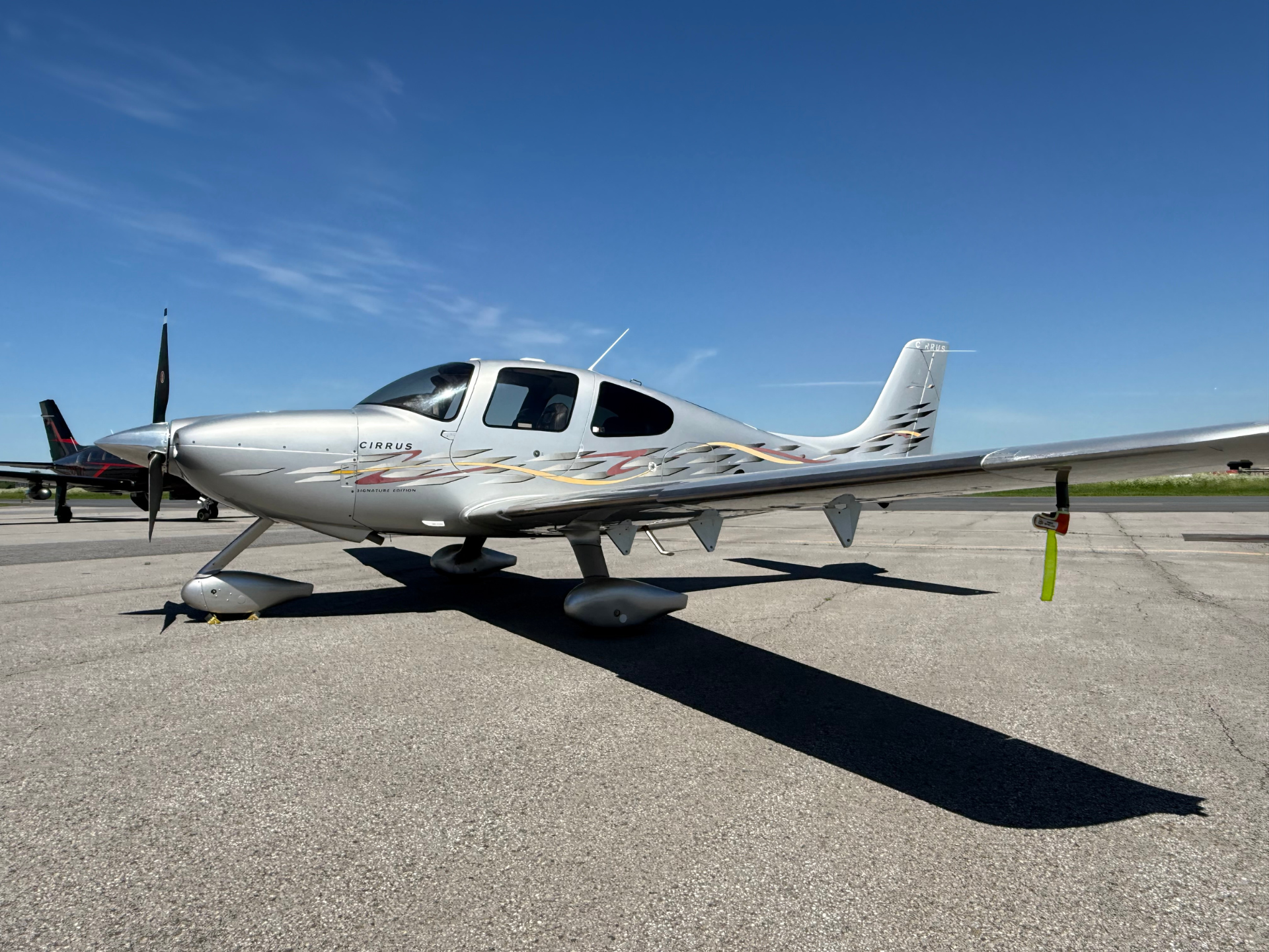 Silver Cirrus single-engine airplane parked on a taxiway with blue sky; colorful ribbon graphics along fuselage and a green propeller at nose.