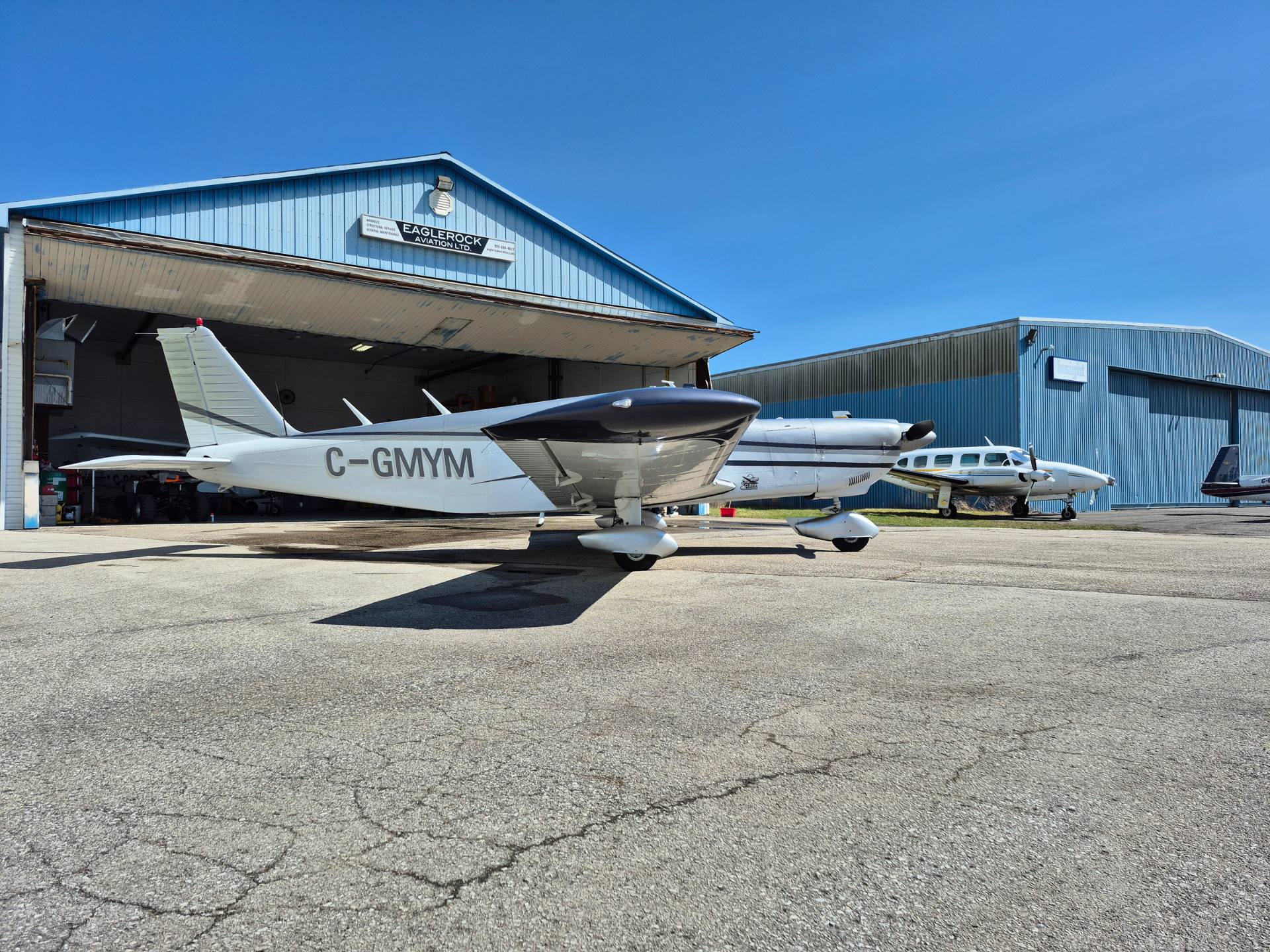 Light single-engine aircraft with registration C-GMYM parked outside a blue airport hangar under a clear blue sky, with another small plane nearby.