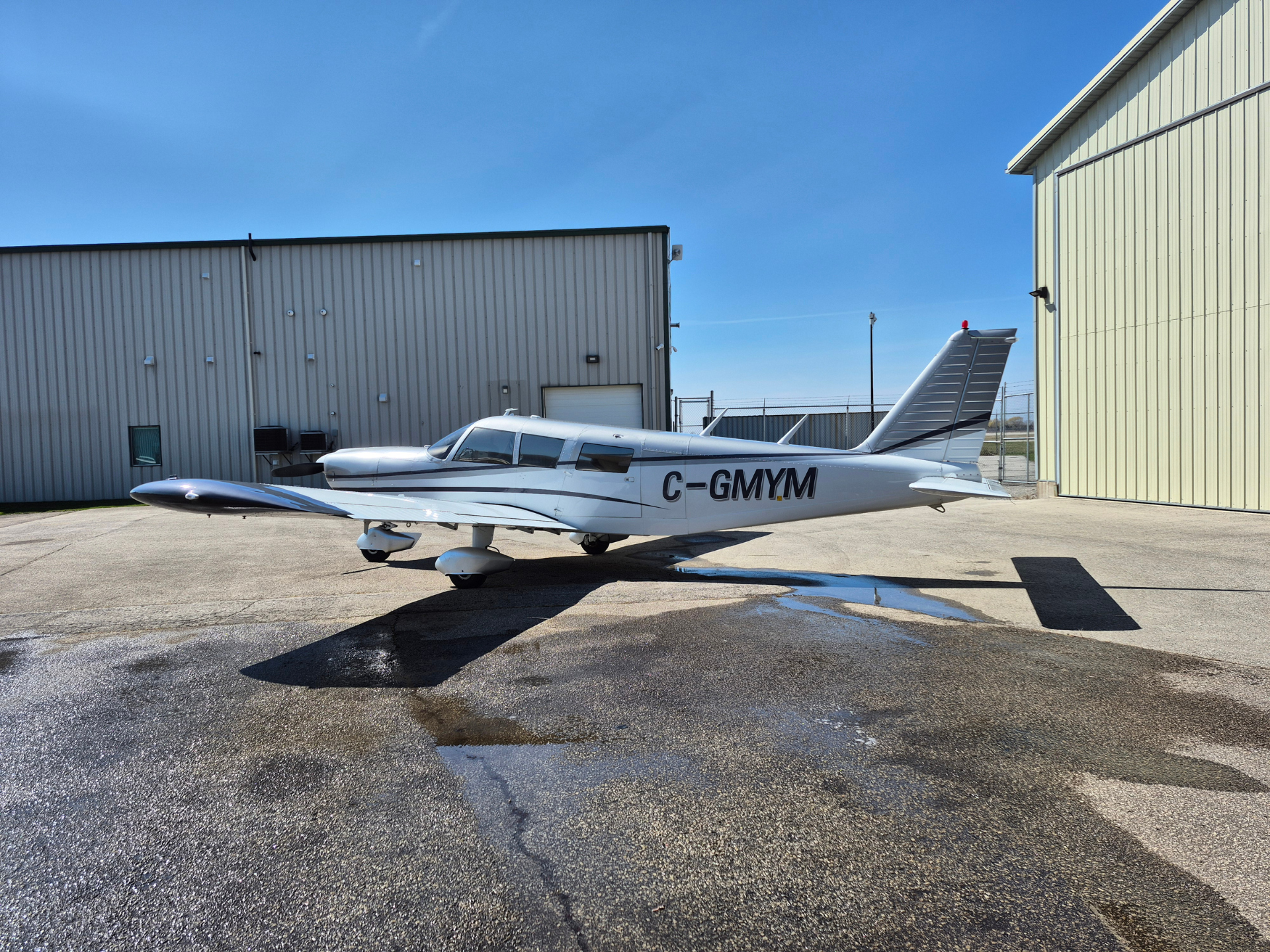 Small white single-engine airplane with registration C-GMYM parked on the tarmac near beige hangars under a clear blue sky.