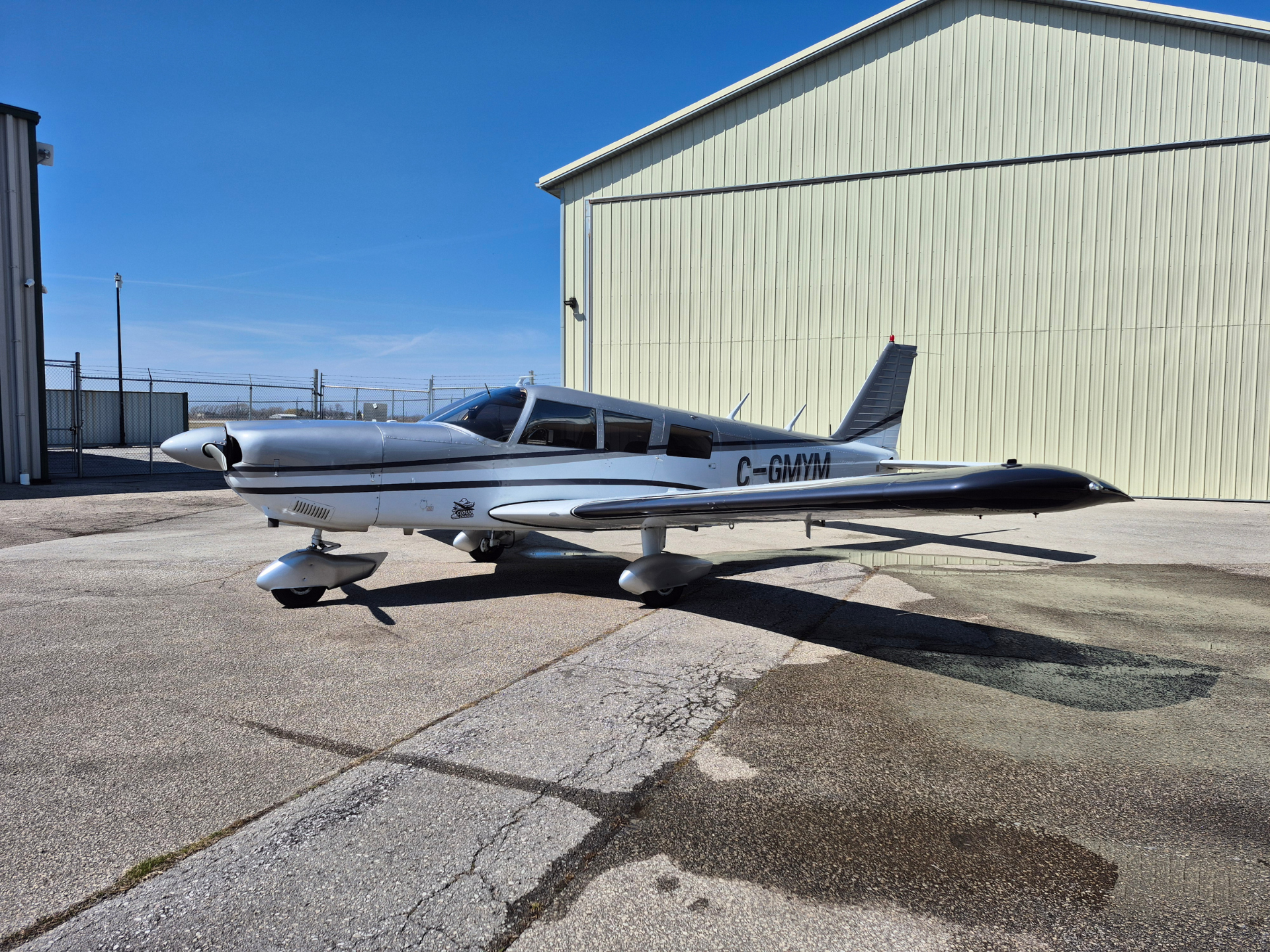 Small silver propeller airplane parked on a concrete apron beside a pale hangar under a bright blue sky, tail number C-GMVN visible