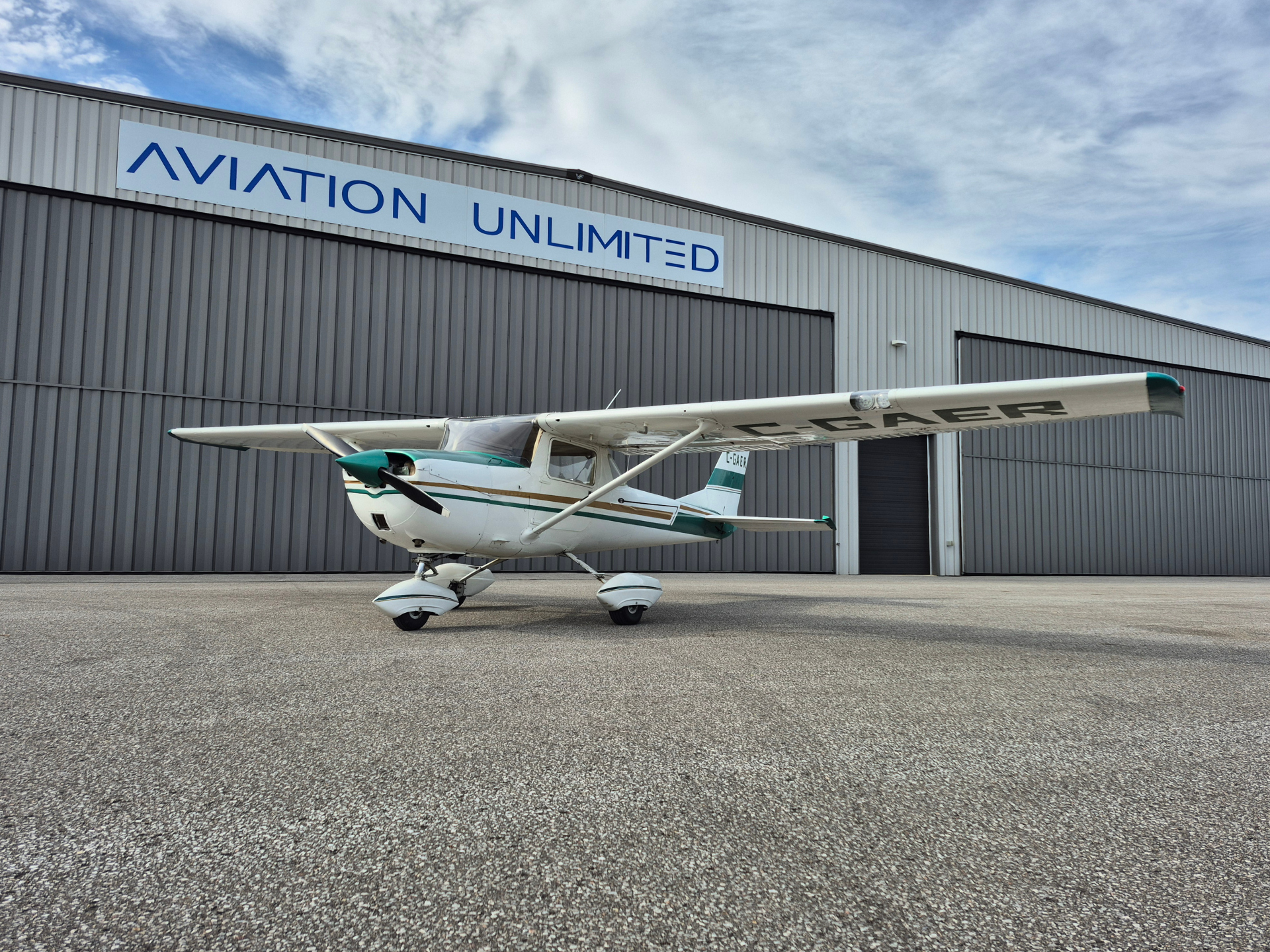 Small white single-engine propeller plane parked outdoors beside a gray hangar with a blue 'AVIATION UNLIMITED' sign above; clear sky.