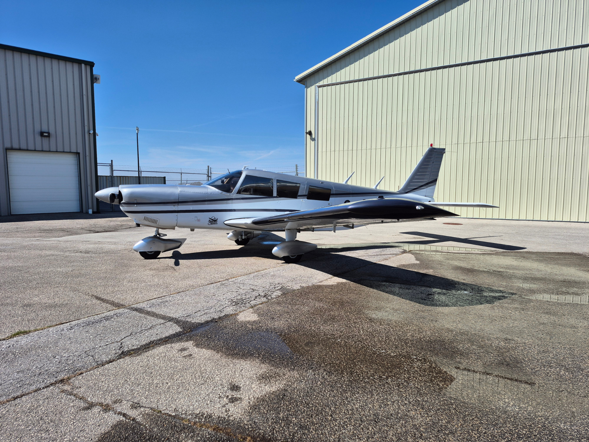 Silver single-engine airplane parked on the apron in front of a beige metal hangar on a sunny day.