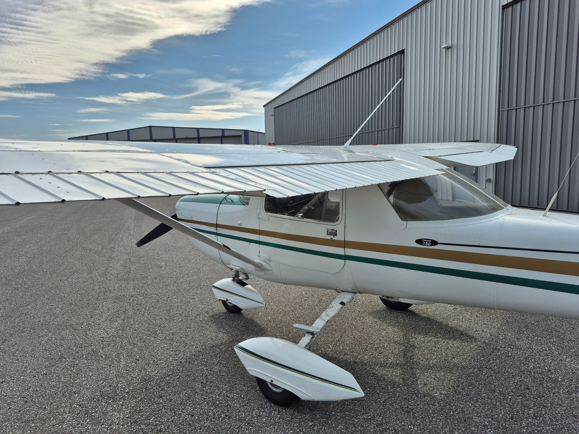 Small white Cessna 150 with green and gold stripe parked on the tarmac beside a corrugated metal hangar, with a wing spanning the foreground.