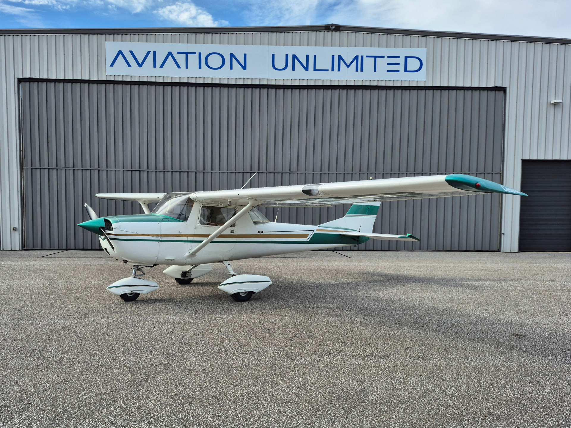 Small single-engine airplane with teal accents parked on a tarmac in front of a gray hangar labeled 'AVIATION UNLIMITED'.