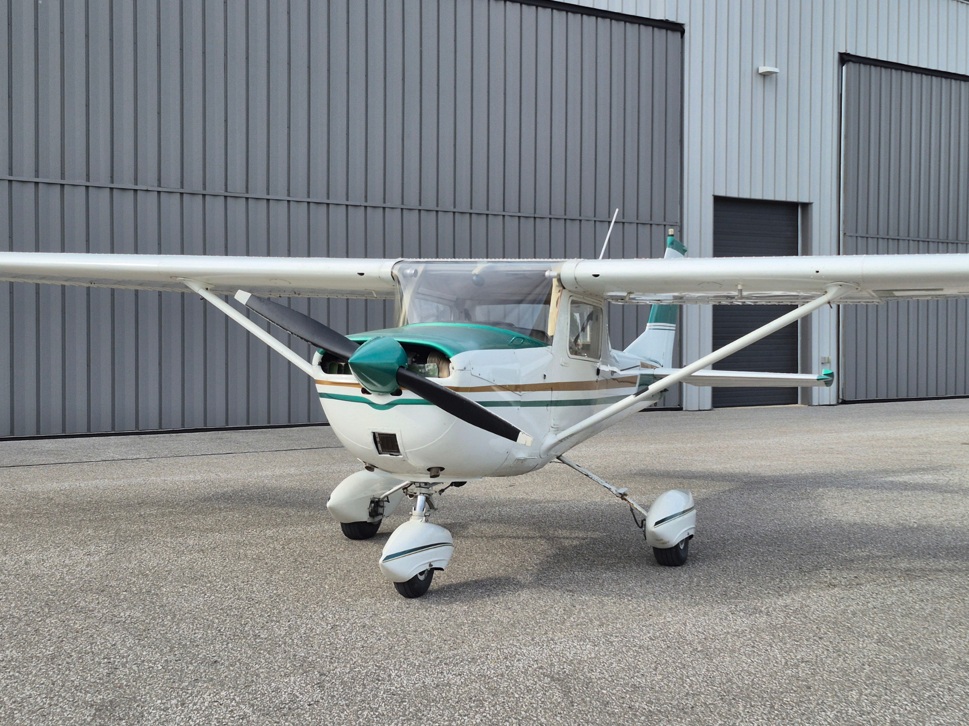 Small propeller airplane with teal and white fuselage parked on the tarmac in front of a gray metal hangar.