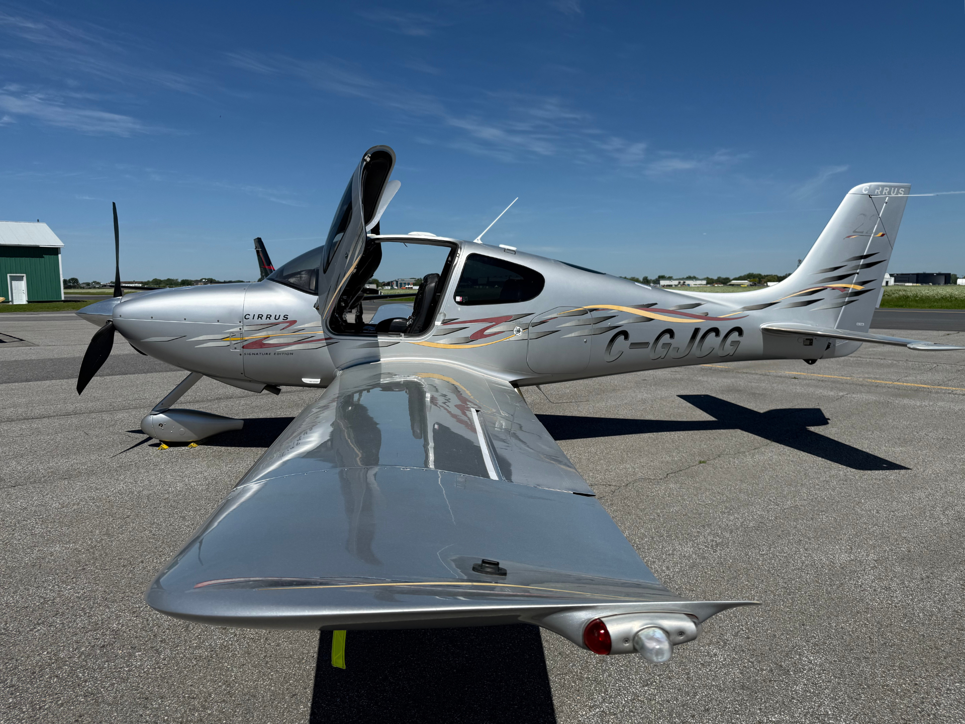 Silver Cirrus small aircraft with open canopy on a sunny airport tarmac, wing toward the camera.