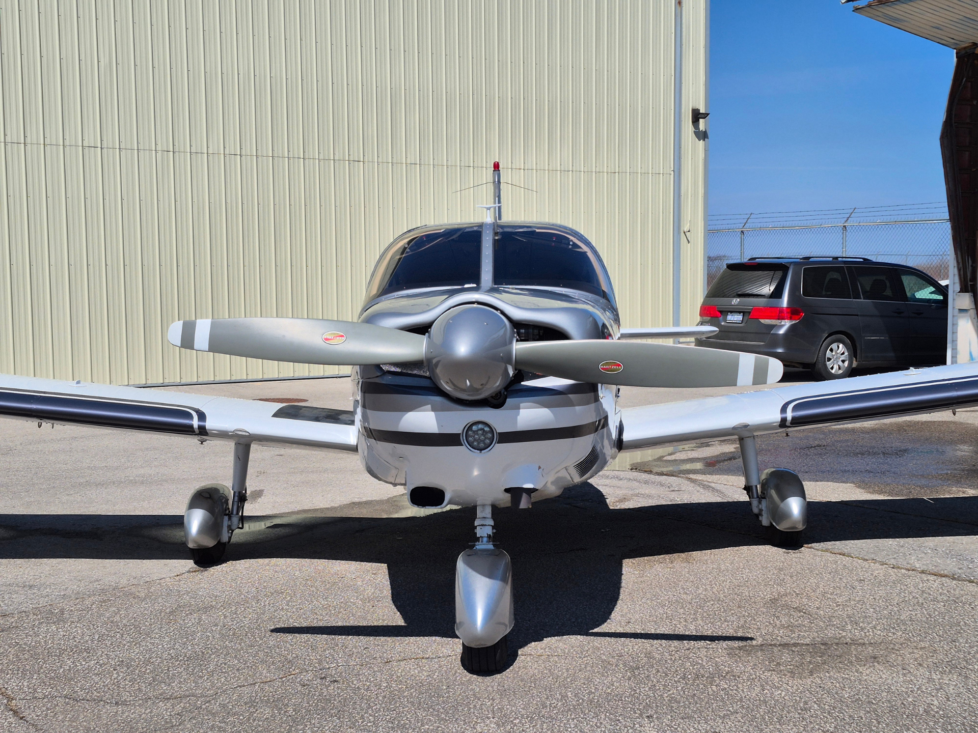 Front view of a small silver single-engine propeller airplane parked on tarmac in front of a beige hangar, with a blue sky and a gray SUV in the background