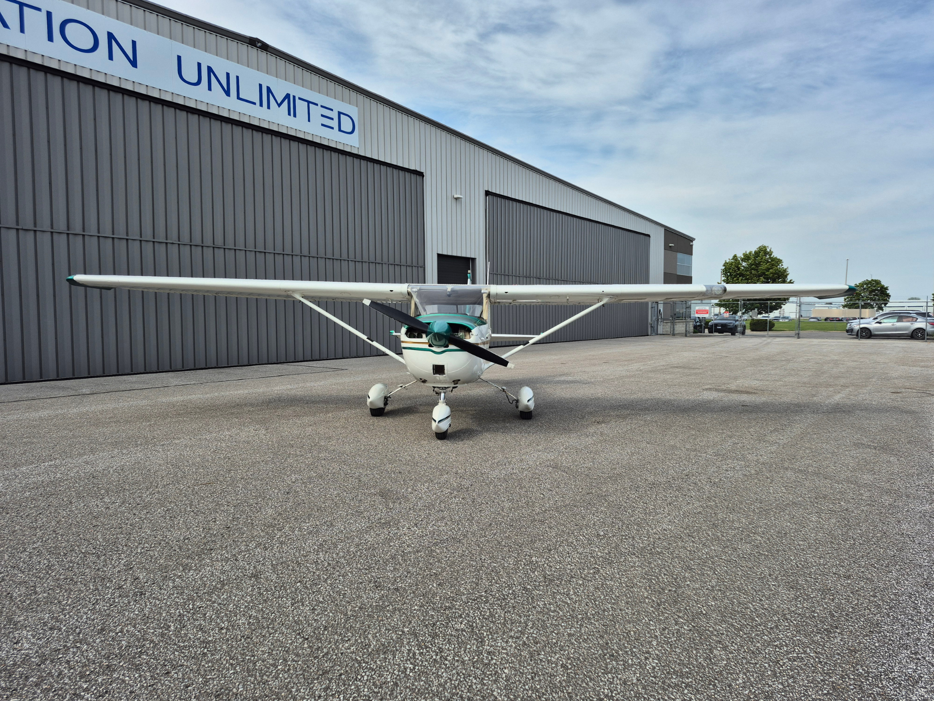 Small white propeller airplane with teal accents parked on an apron in front of a gray hangar. A large blue 'UNLIMITED' sign is visible on the building.