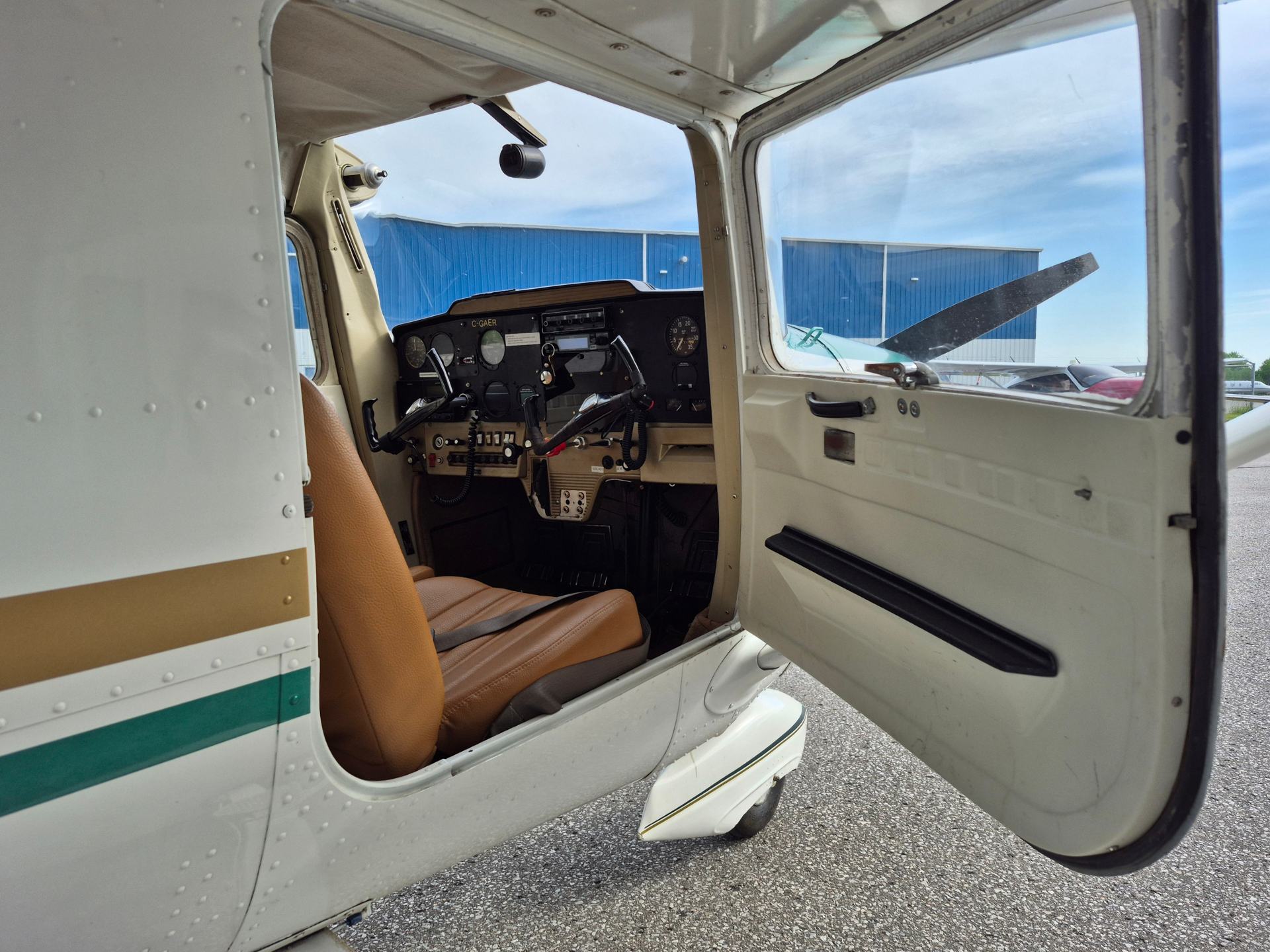 Open door of a small aircraft showing the cockpit with dials and controls, tan seat, and a propeller outside against a blue hangar.