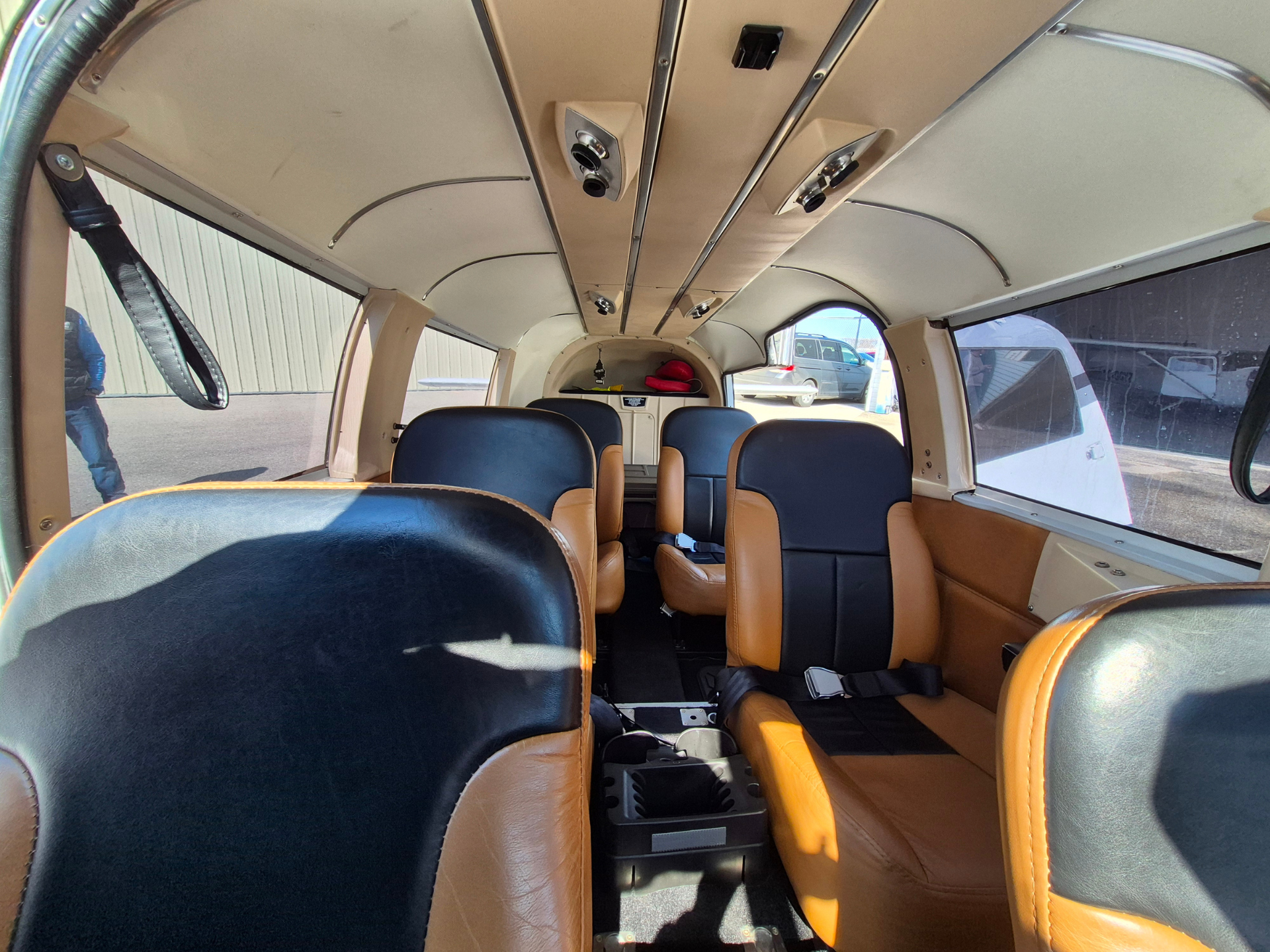 Interior of a small airplane cabin with tan and dark blue seats, passenger belts visible, sunlit windows to the right and back view of cockpit area ahead