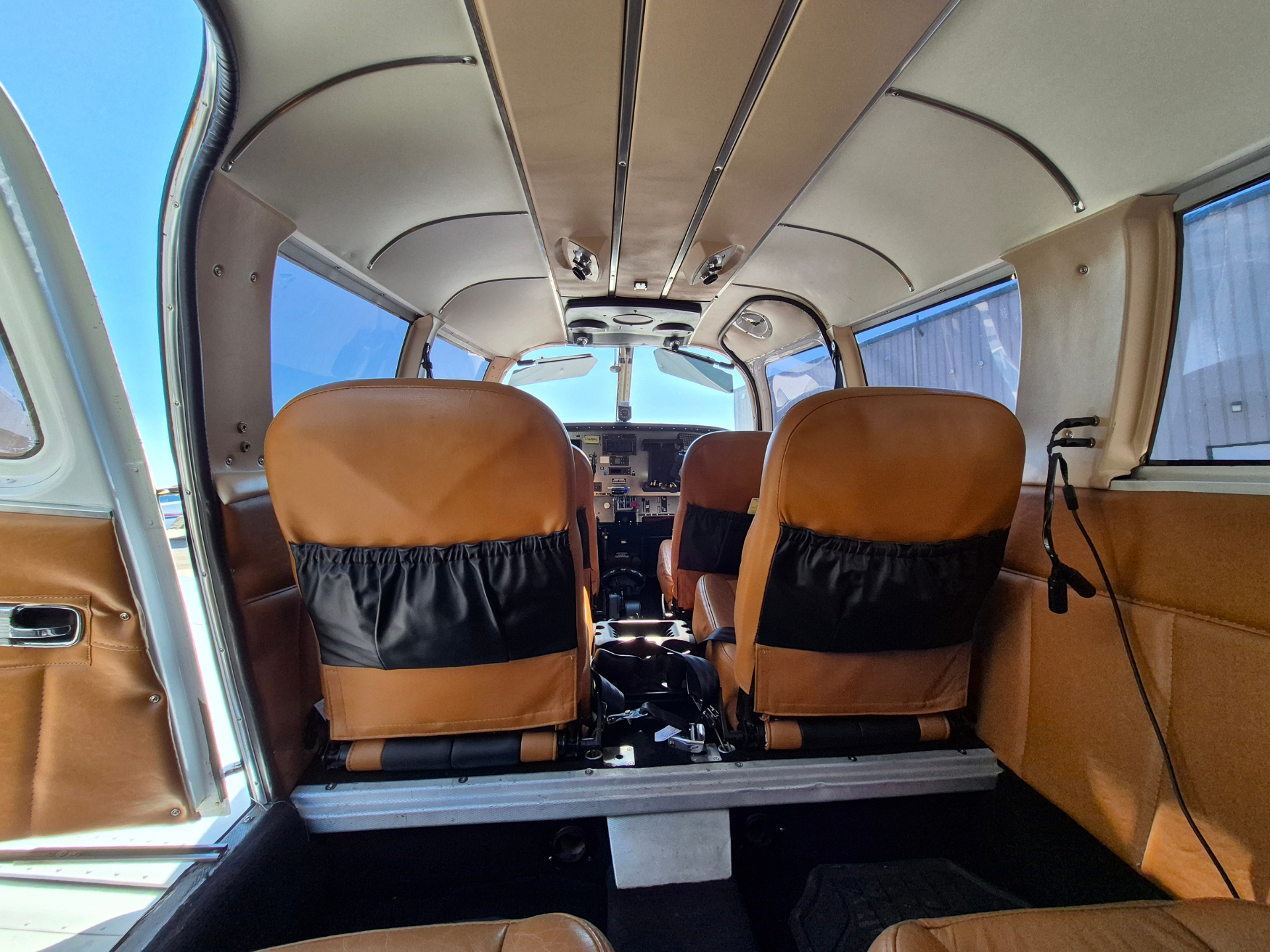 Interior of small airplane cabin with tan seats, cockpit controls visible at the front, blue sky outside the side windows.