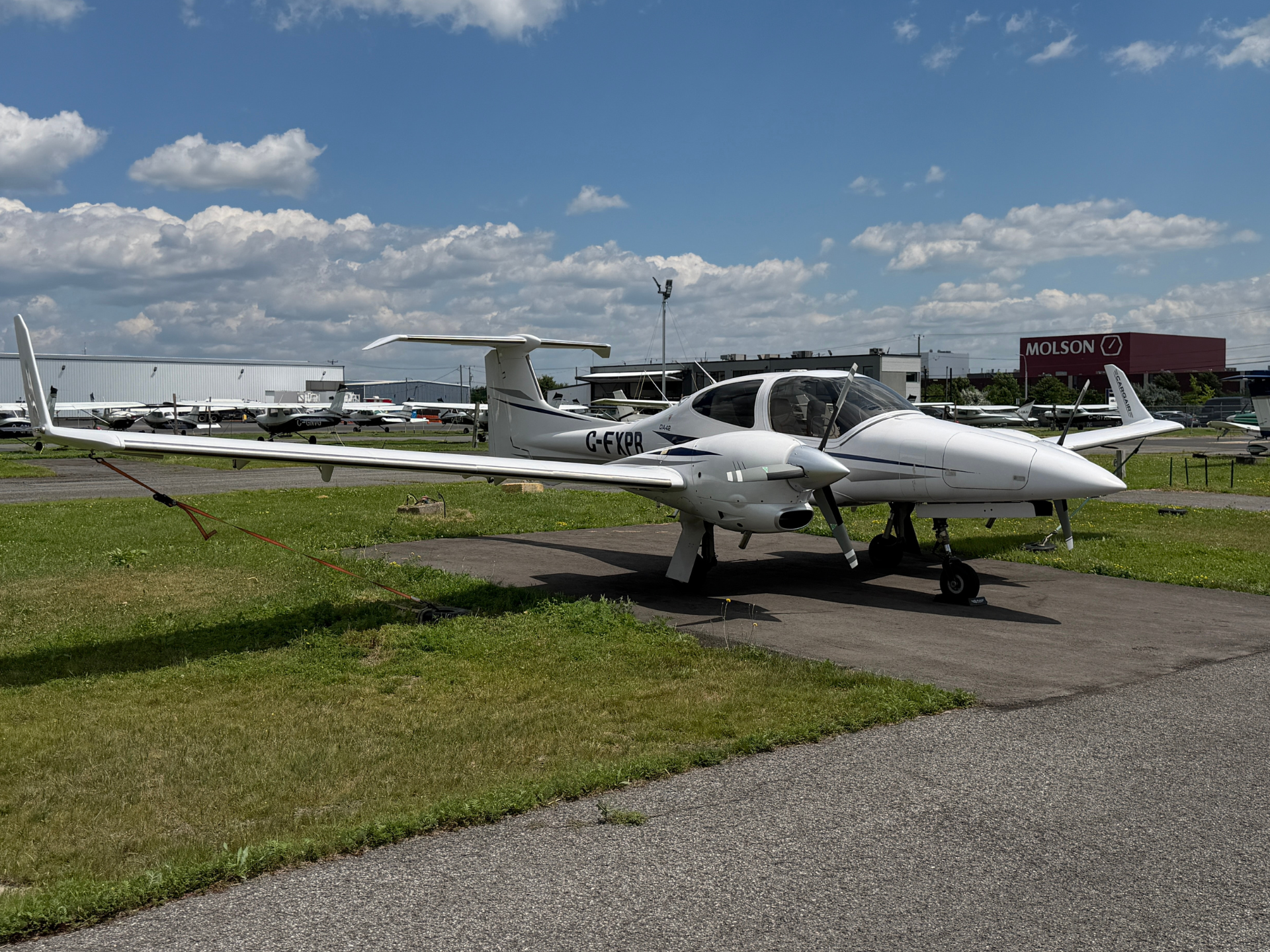 White Diamond DA42 light aircraft parked on a tarmac at a small airport, with other planes, grass, and a Molson building in the background.