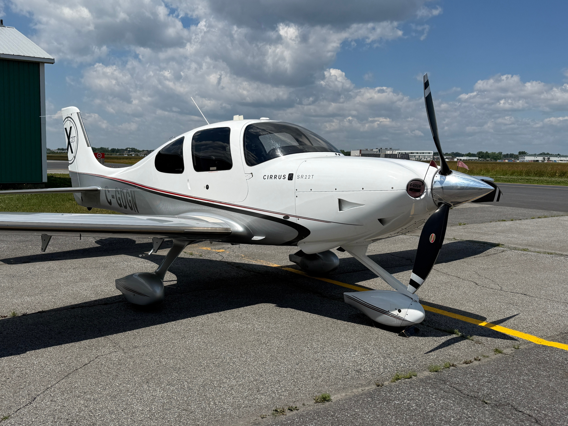 White Cirrus SR22T aircraft parked on the tarmac beside a green hangar, under a partly cloudy blue sky.