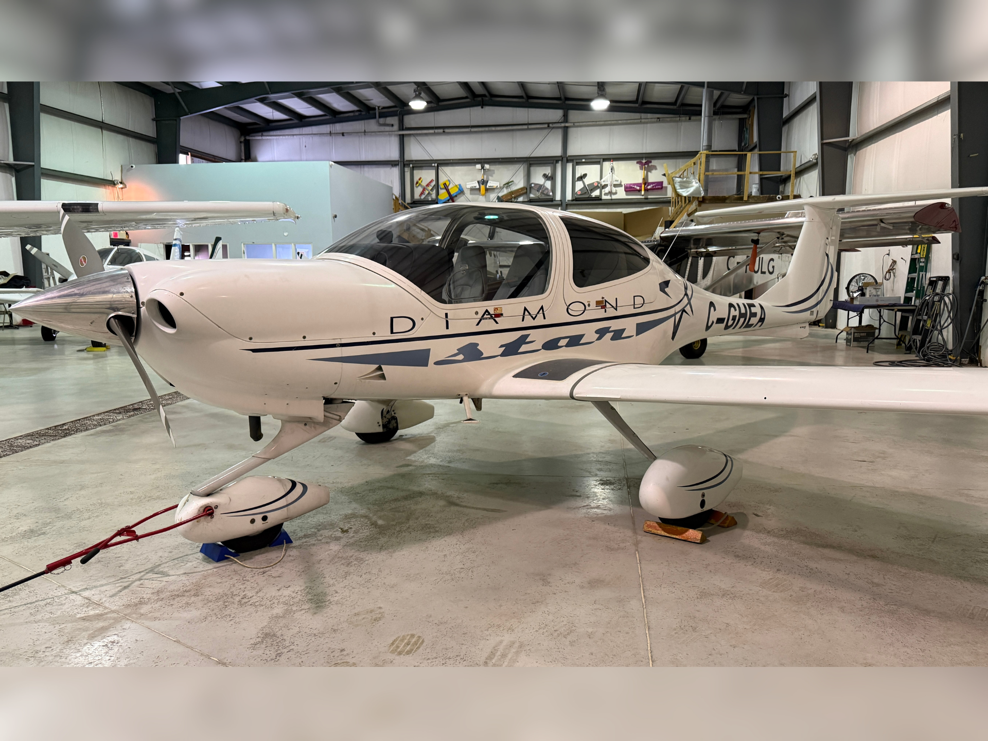 White Diamond Star airplane in a hangar, cockpit canopy closed, with workshop tools and other aircraft in the background.