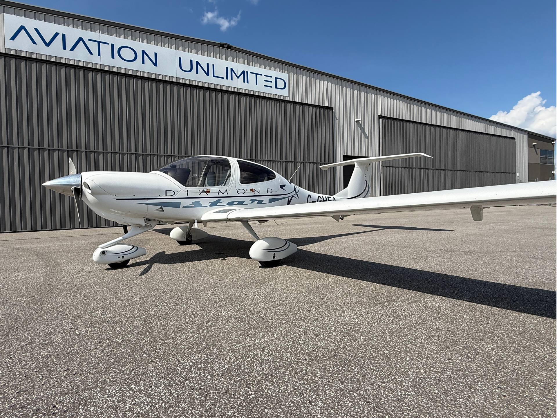 White single-engine airplane parked on the tarmac in front of a gray hangar with a blue 'AVIATION UNLIMITED' sign under a clear blue sky.