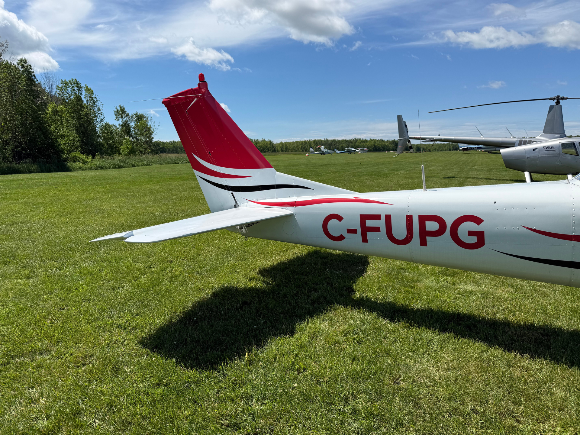 Tail and rear fuselage of a small white airplane with red tail and black stripe, parked on a grassy airfield; other planes visible in the background