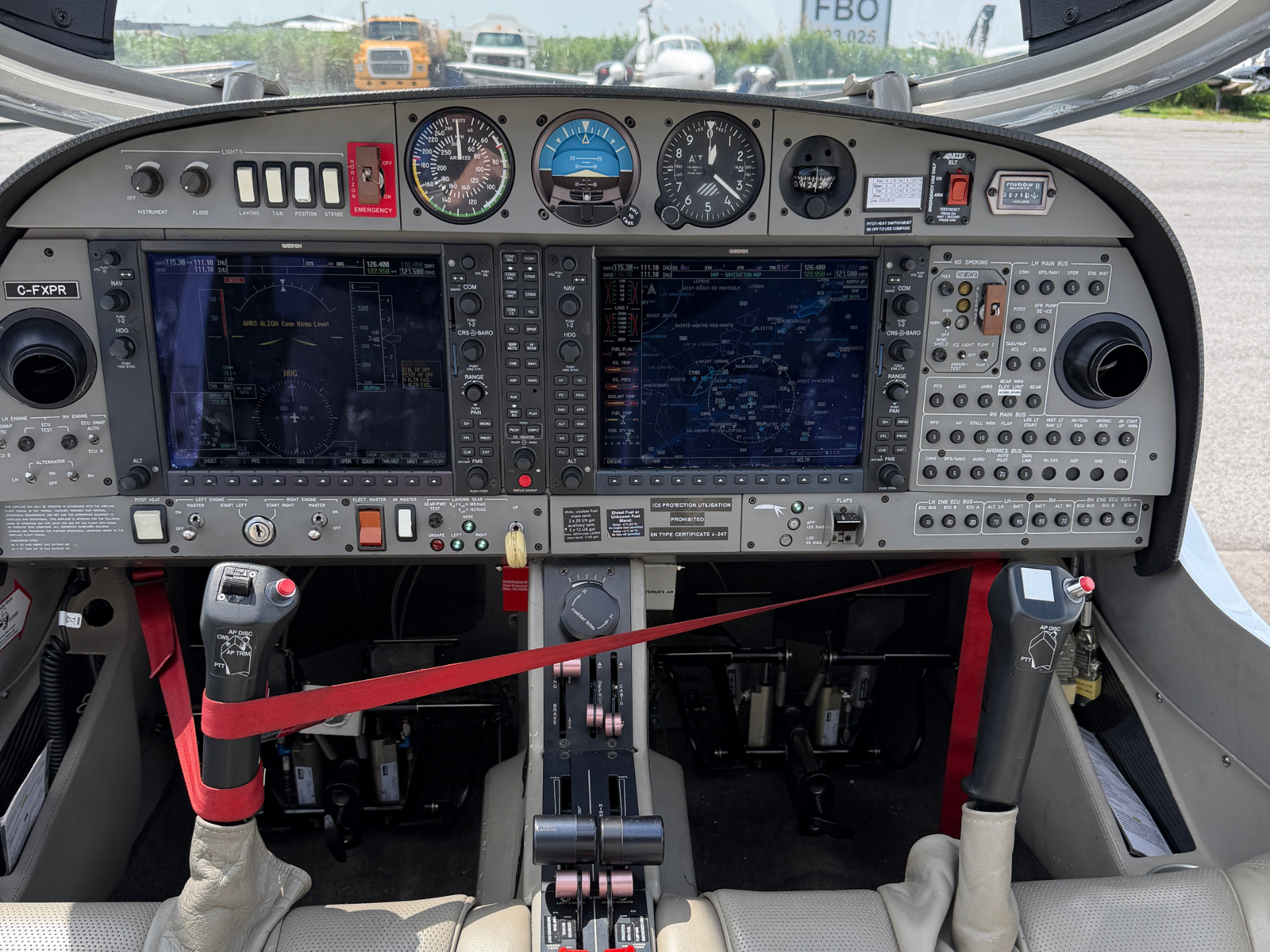 Front cockpit panel of a small airplane showing dual large flight displays, analog gauges, and red safety harnesses at the seats, with throttles in the center.