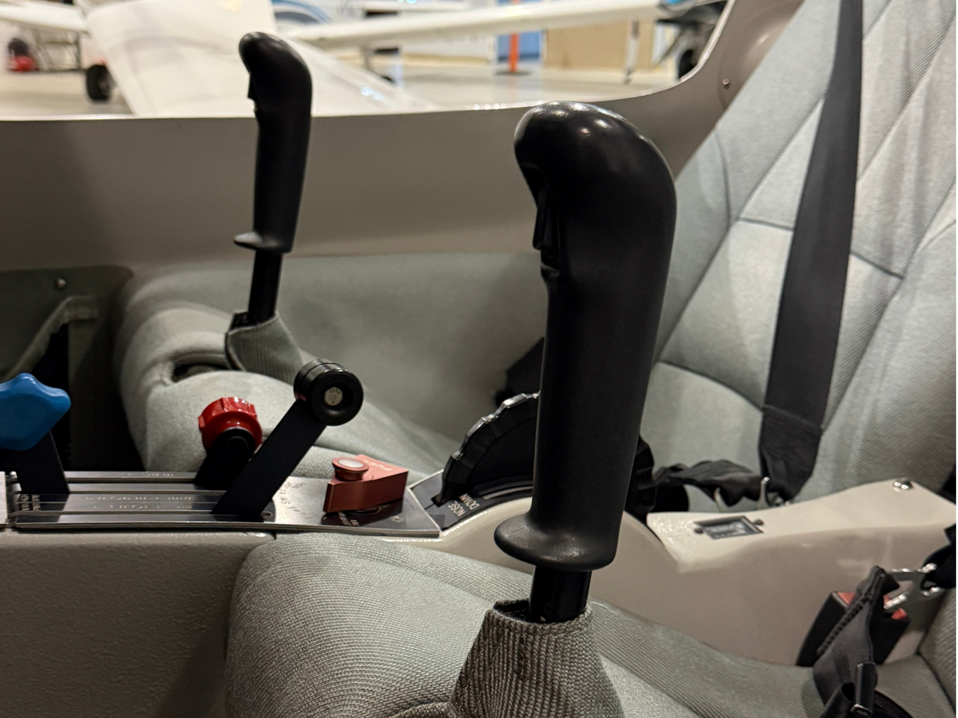 Close-up of airplane cockpit controls: black joystick levers, red knobs, and gray upholstery in view of the pilot area.